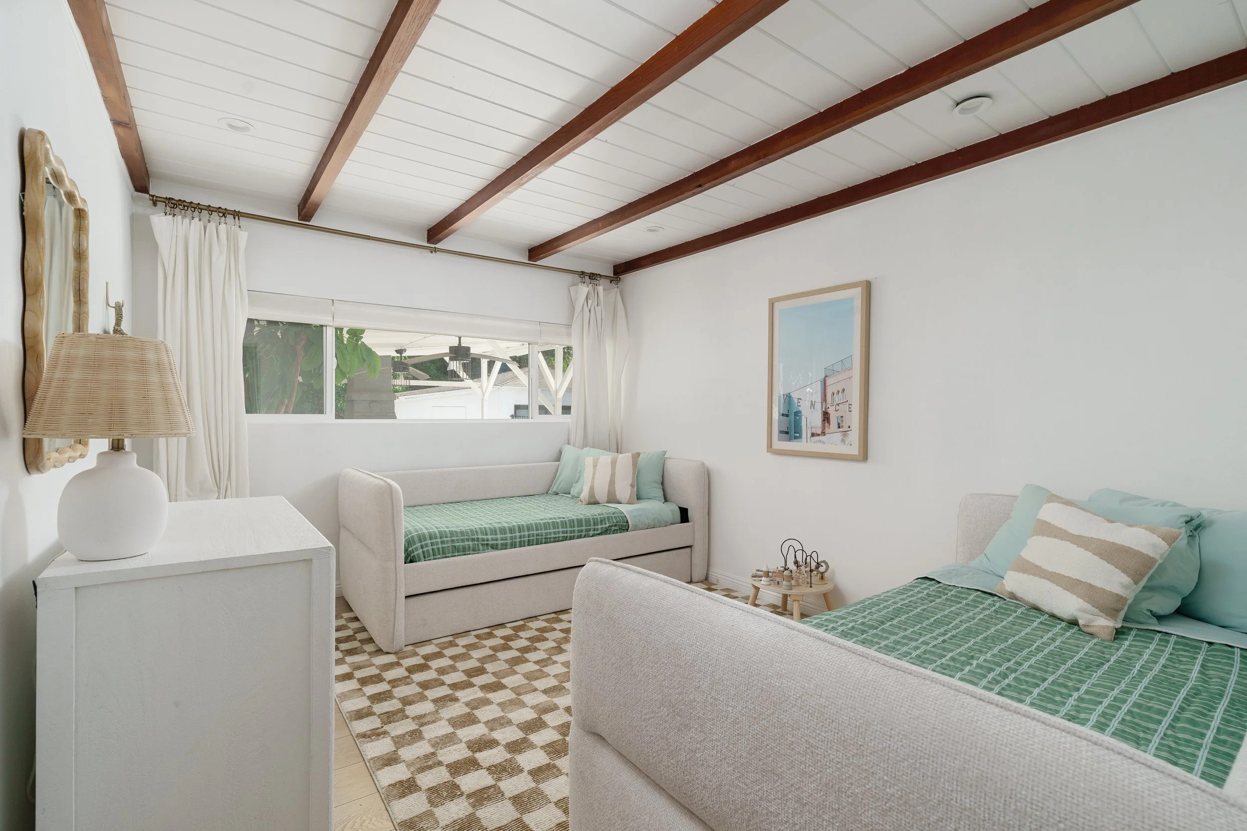 A bright bedroom with white walls and ceiling, wooden beams, and two twin beds with green bedding, white pillows, and beige headboards. A white side table with a lamp is next to the beds, and there's a window with curtains showing greenery outside. A framed picture decorates the wall.