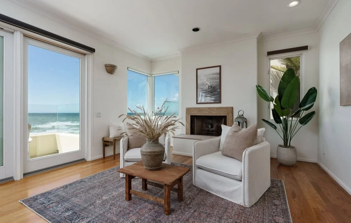 Living room with large windows overlooking the beach, white armchairs, a fireplace, potted plant, and coastal decor.