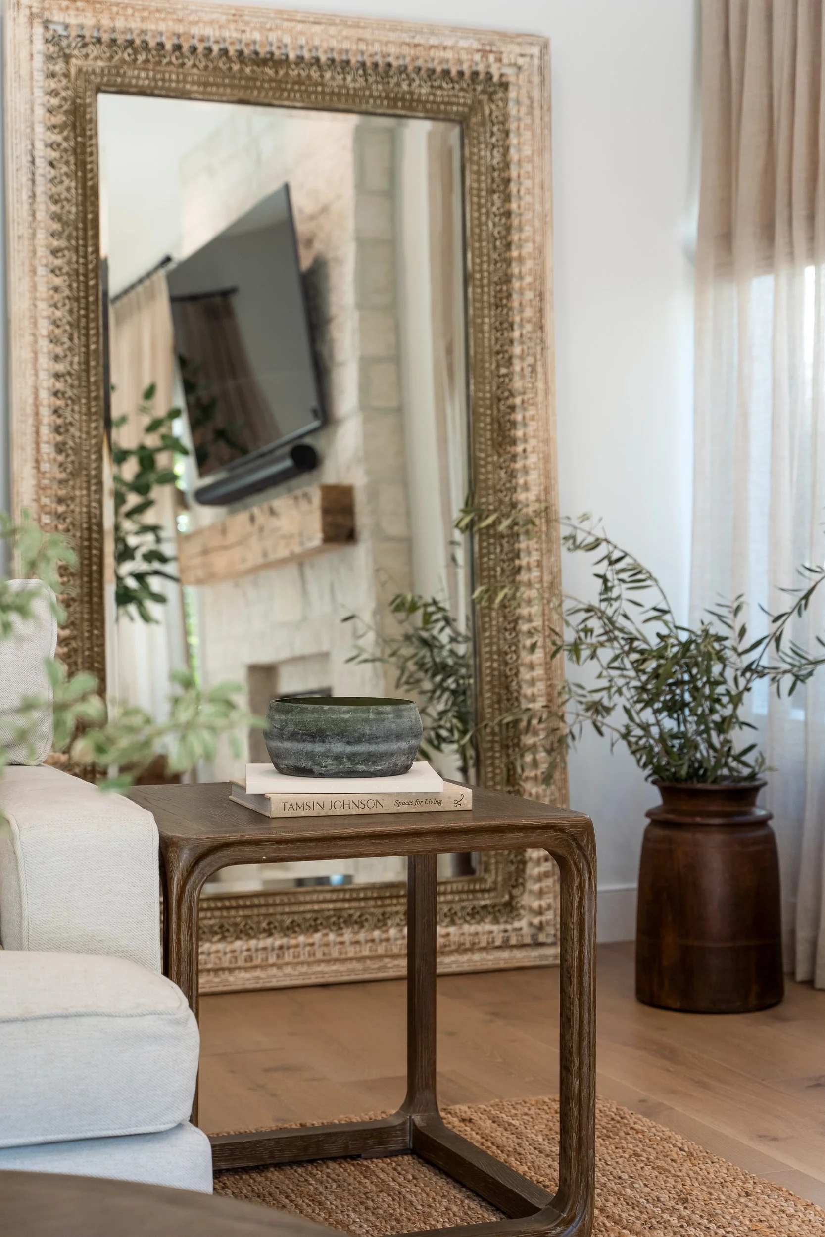 A living room corner with a large decorative mirror, a beige sofa, a wooden side table with a stone bowl and books, a potted plant in a dark wooden container, and a wall-mounted TV.