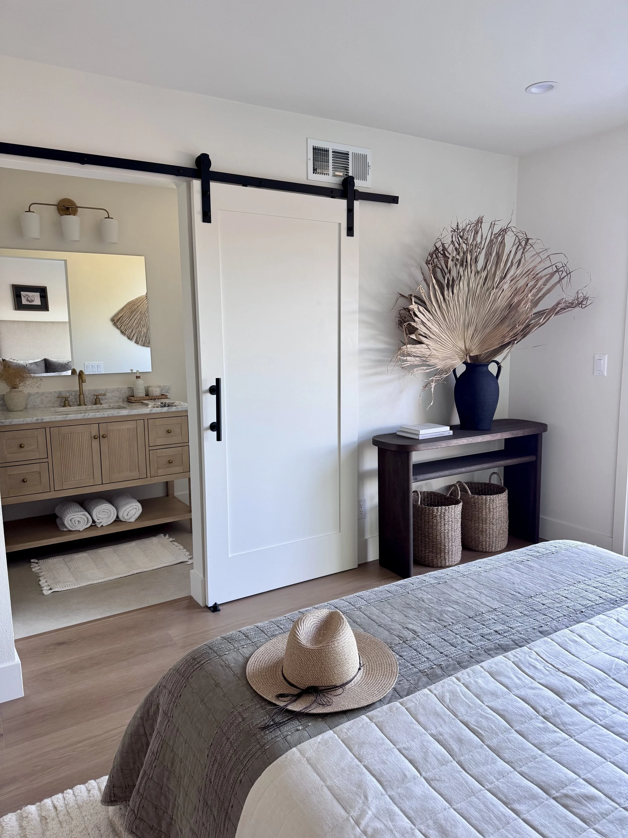 A bedroom with a bed, hat, and a dark wooden console table holding a large vase with dried palm leaves. A sliding barn door on a black track leads to a bathroom, and there is a large mirror above a wooden vanity in the bathroom. Wicker baskets are on the floor of the console table, and the room has white walls and wood flooring.