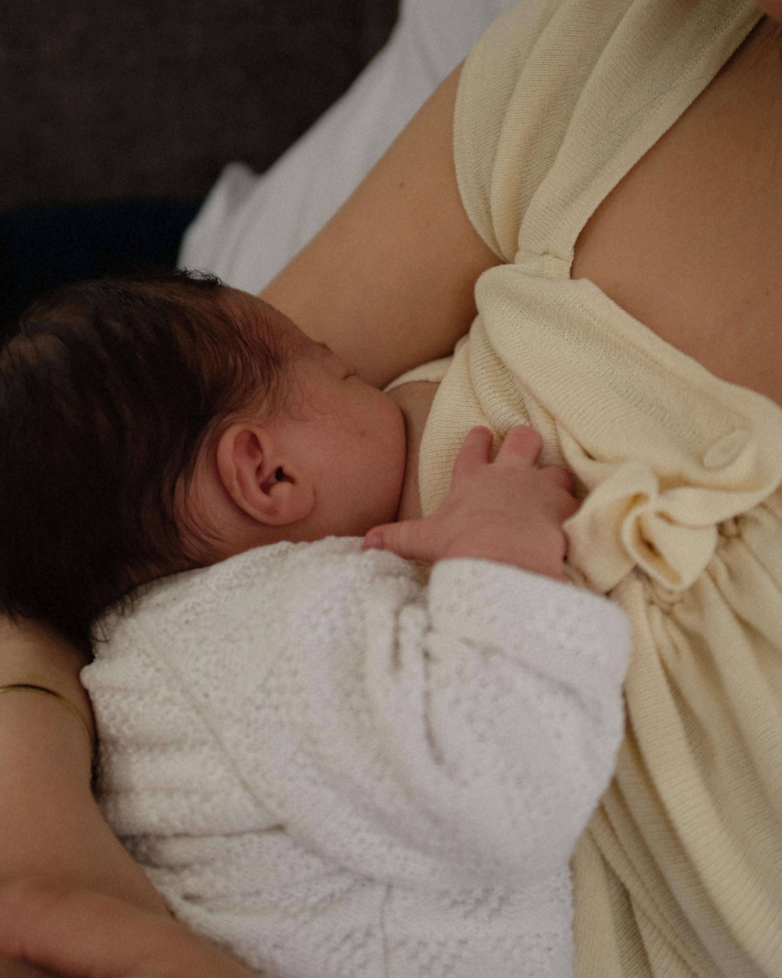 Close-up image of a newborn baby breastfeeding while lying on a white blanket, wearing a light-colored onesie, on a dark surface.