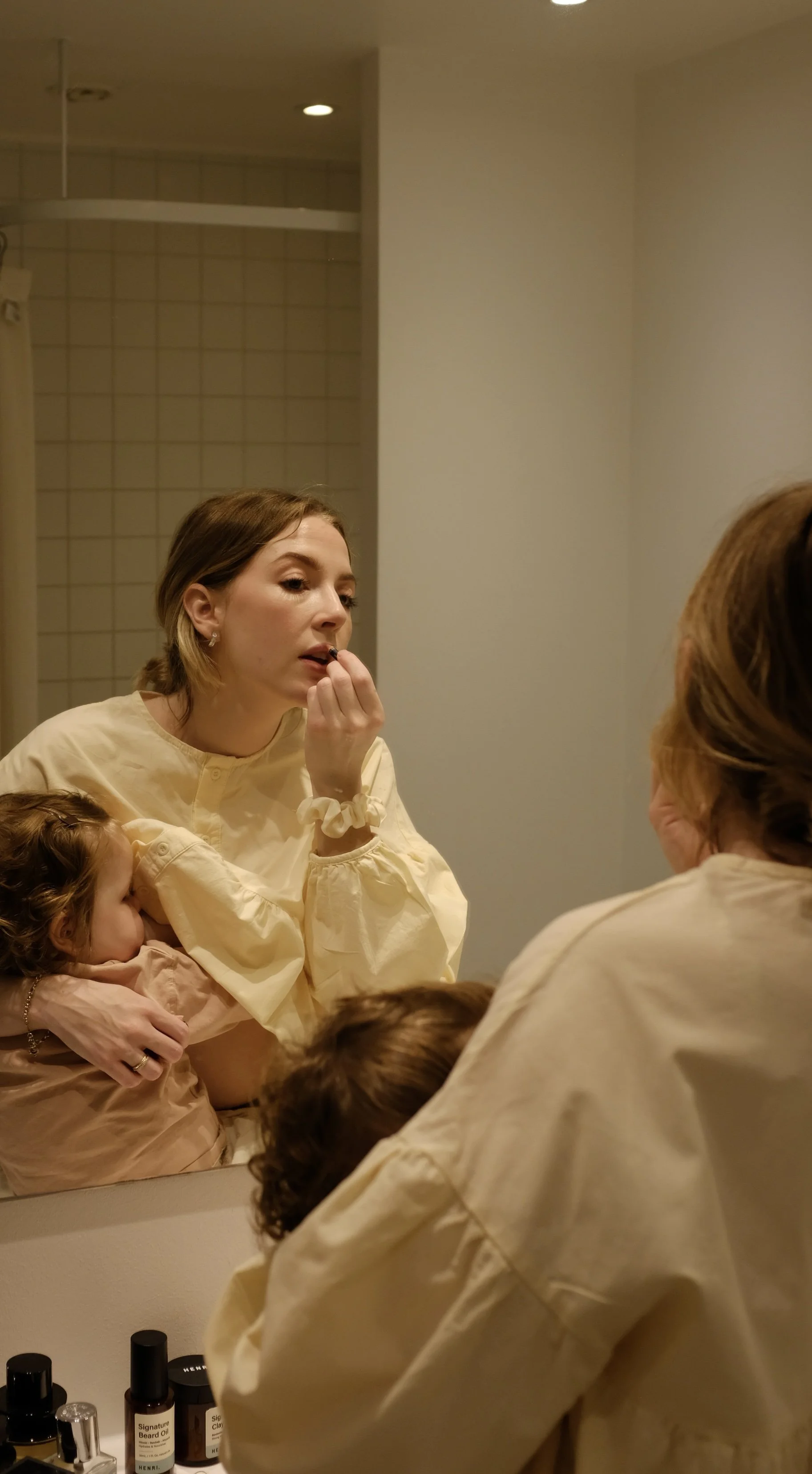 A woman applying lipstick while holding a child, seen in a mirror in a bathroom.