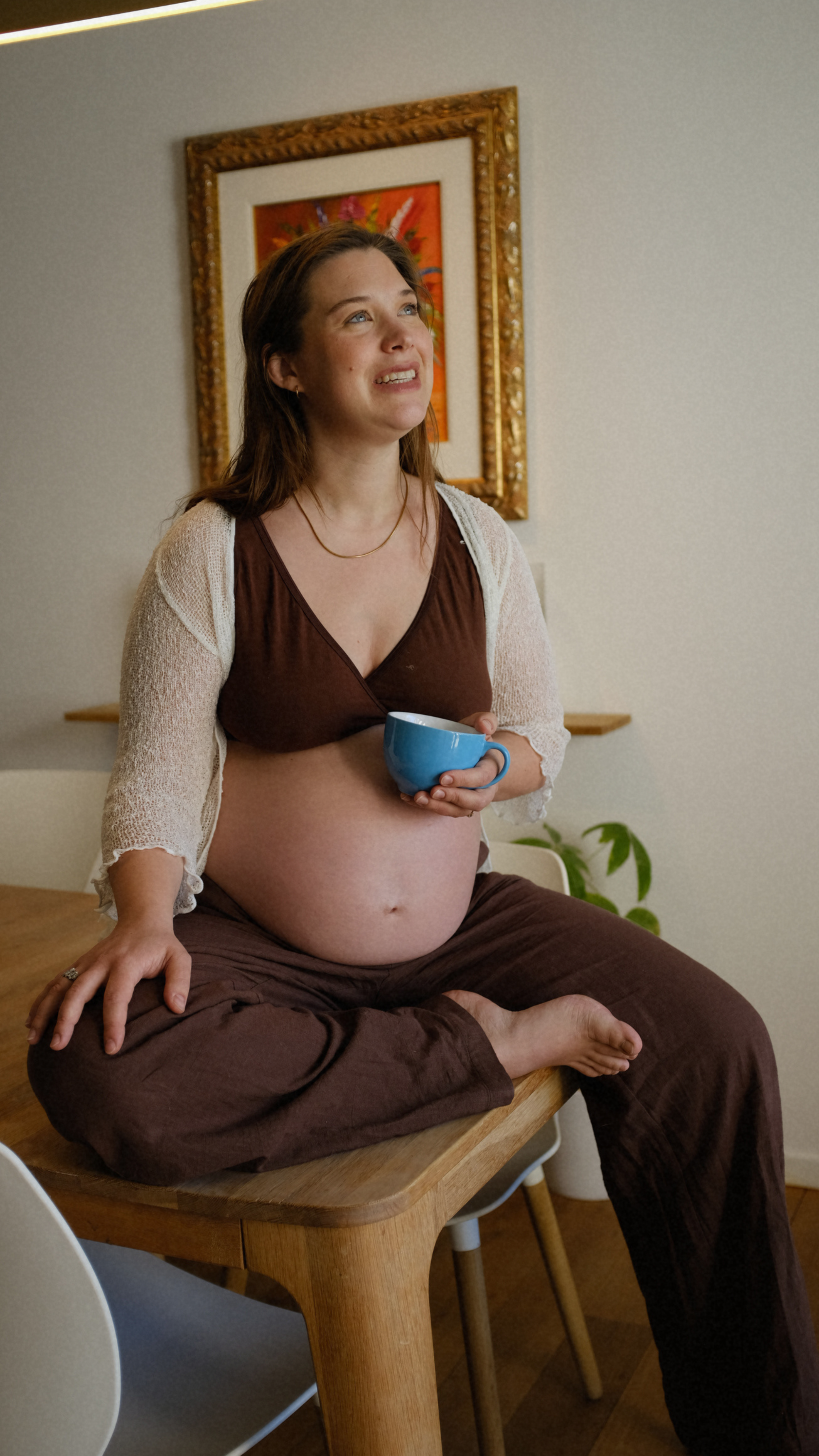 A pregnant woman sitting cross-legged on a wooden table, holding a blue mug, in a room with a framed colorful art piece and a green plant in the background.