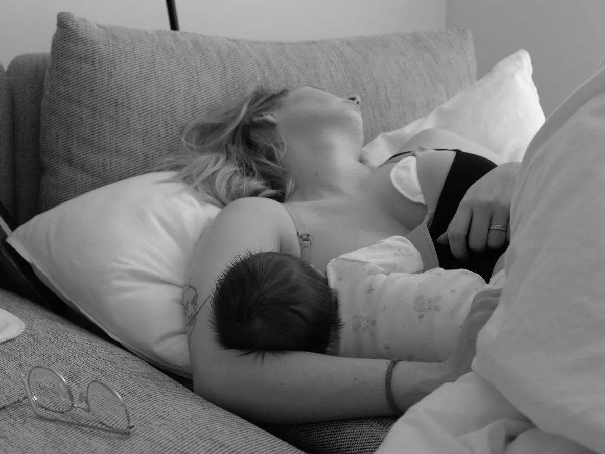 A woman lying on a bed, breastfeeding a baby while sleeping, with glasses resting on the bed nearby.