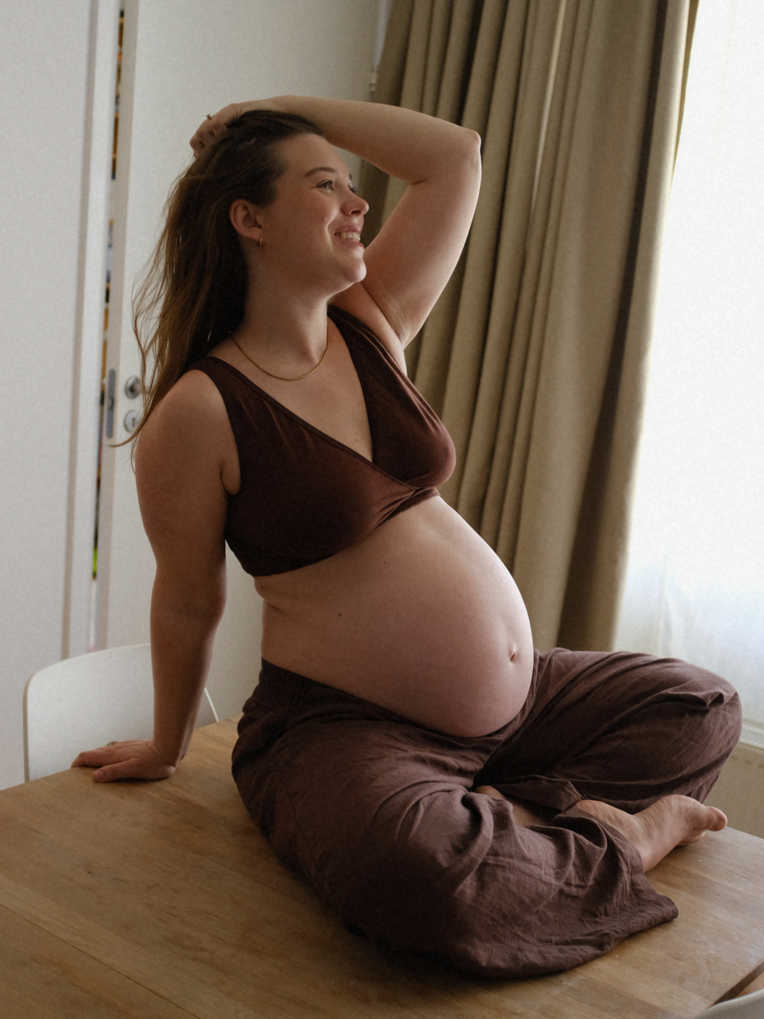 Pregnant woman with long brown hair in a brown crop top and loose brown pants, sitting on a wooden table, smiling with one arm resting on her head and the other hand on the table, near a window with beige curtains.