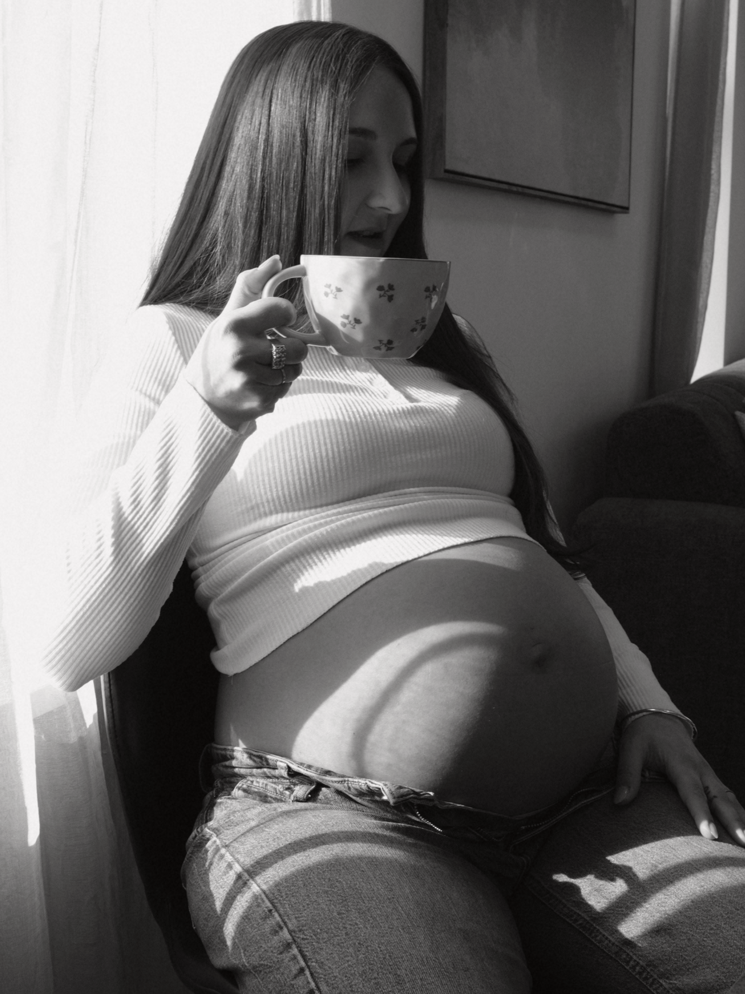 A woman with long dark hair sitting against a wall, holding a floral mug near her face, wearing a long-sleeved shirt and jeans.