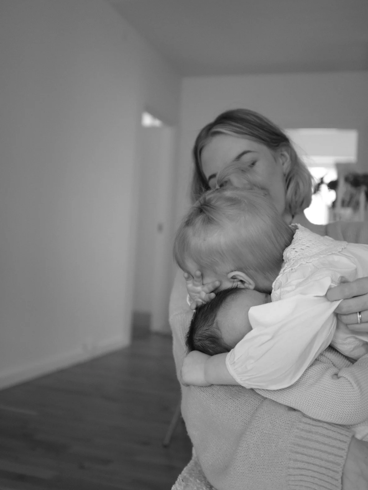 A woman embraces two young children, one with blonde hair and the other with darker hair, in a comforting hug indoors.