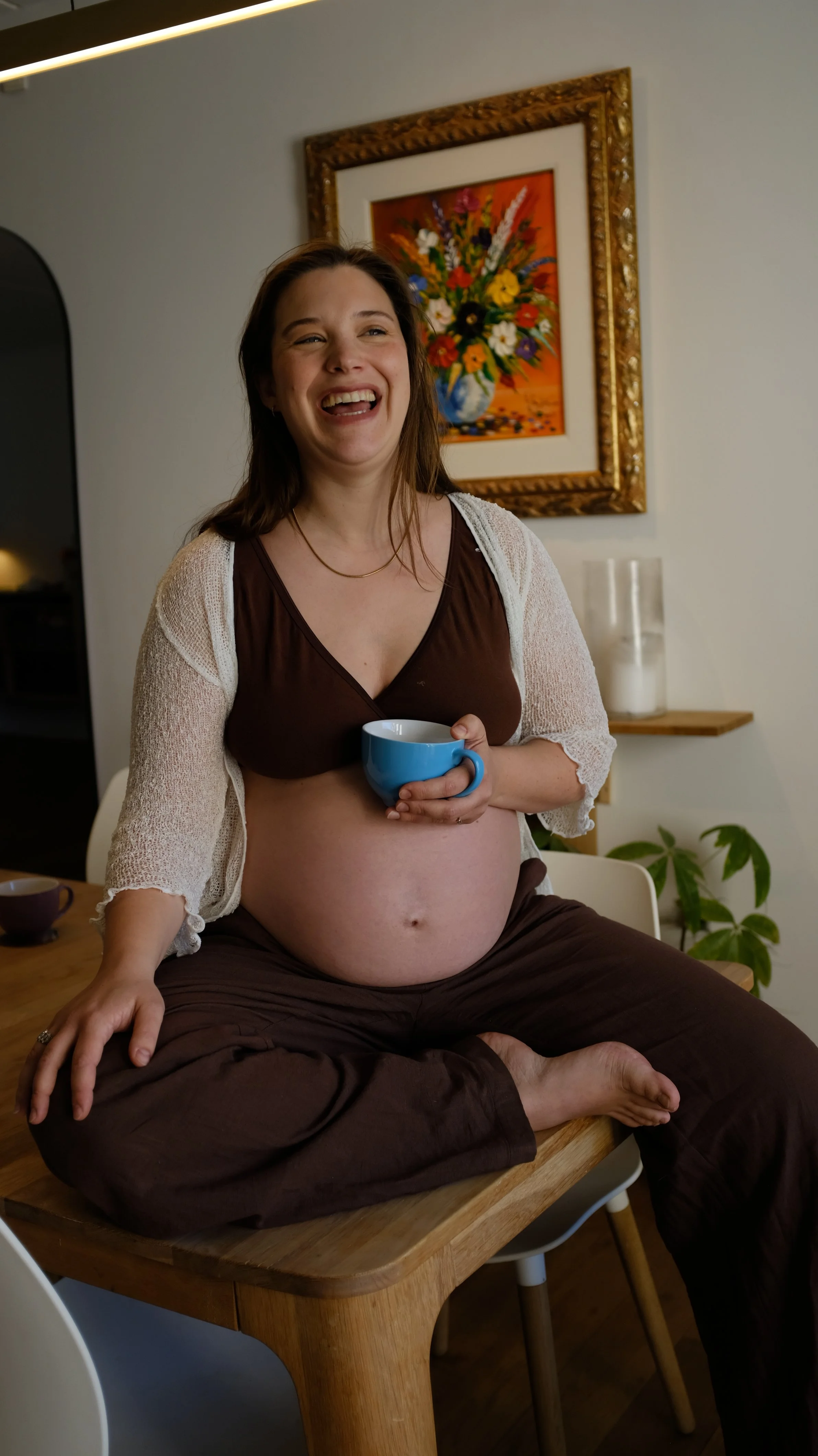 A pregnant woman sitting on a wooden table, holding a blue mug, smiling and laughing, with a painting of flowers on the wall behind her, and plants nearby.
