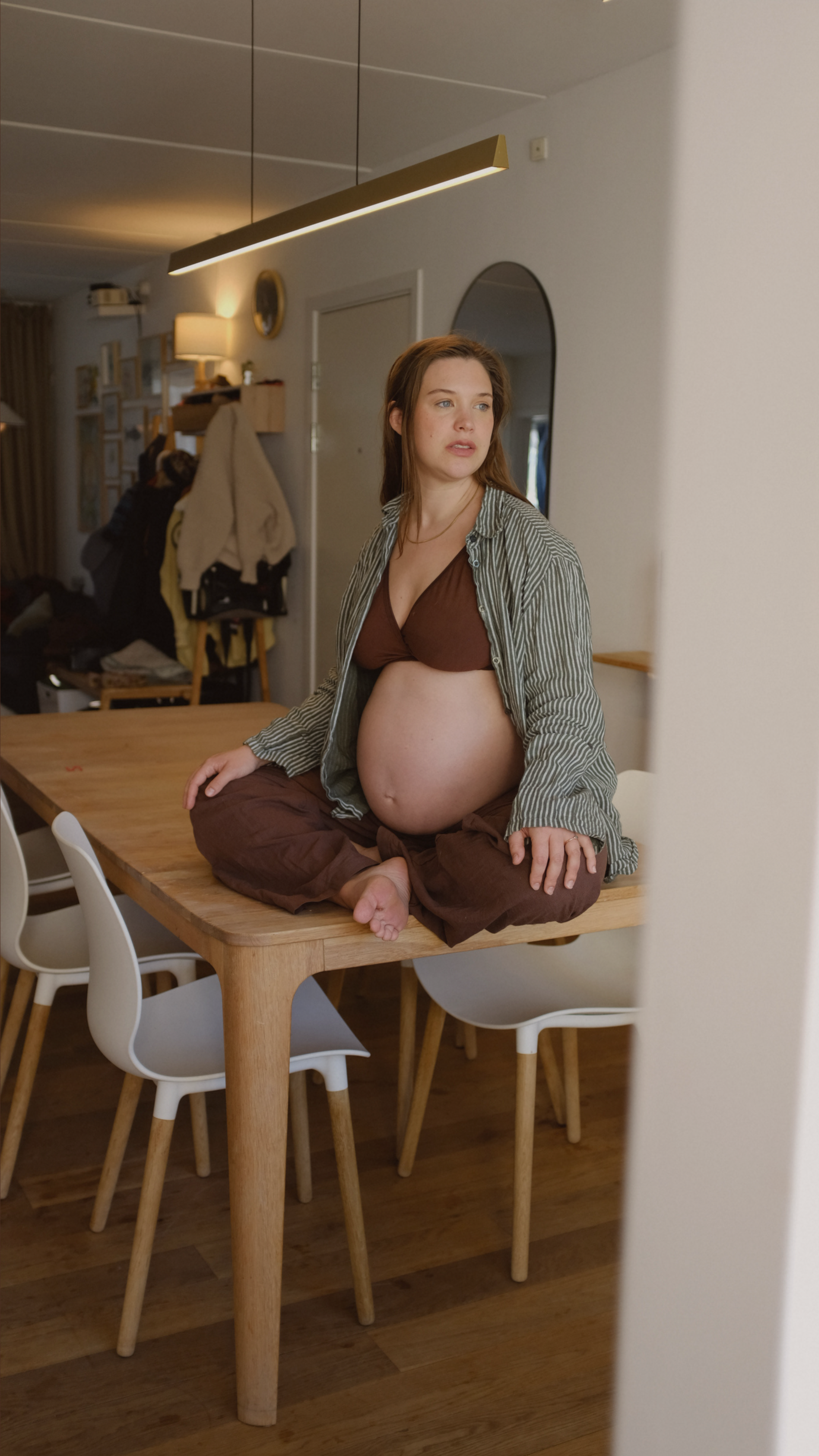 A pregnant woman sitting cross-legged on a wooden dining table, wearing a brown bra and an unbuttoned shirt, with a focused expression, in a modern kitchen or dining area.