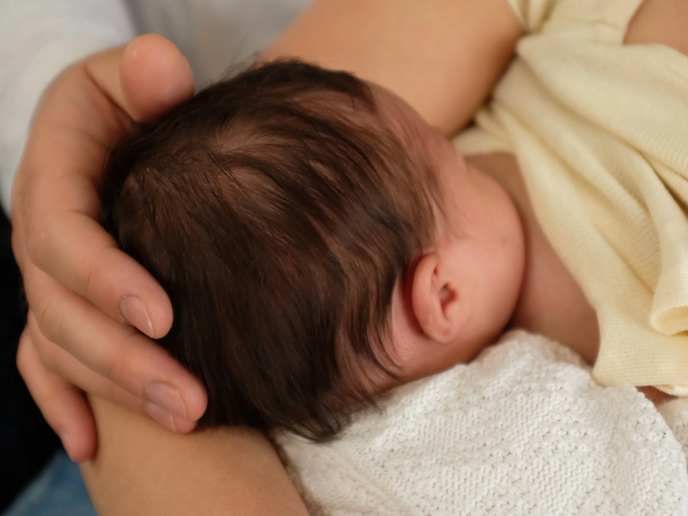 Close-up of a hand gently holding a baby's head with dark hair, with the baby lying on a soft surface and wearing a yellow garment.