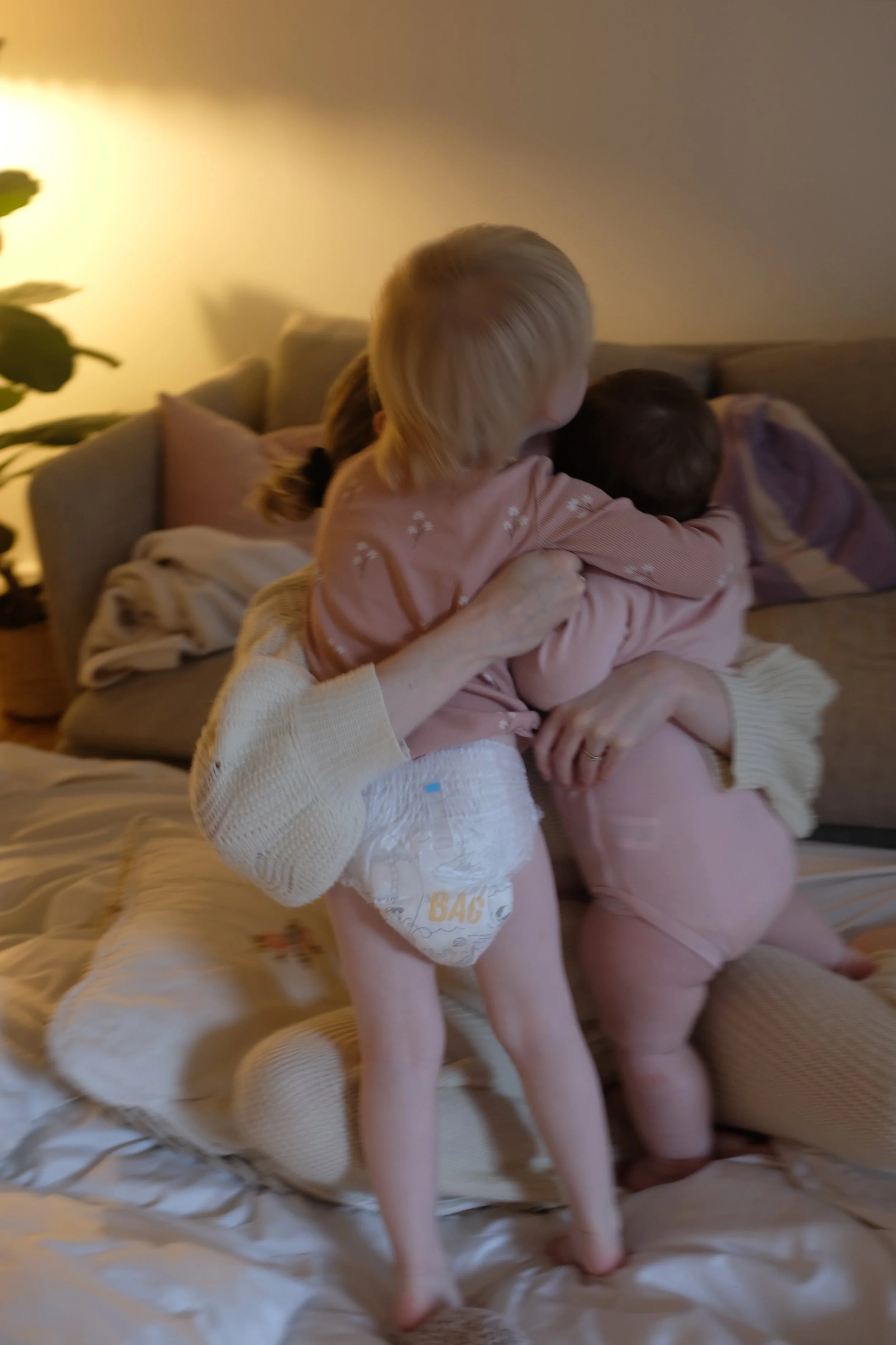 Two young children, a girl and a boy, hugging on a beige couch in a warmly lit living room. The girl has blonde hair and is wearing a pink shirt. The boy has dark hair and is wearing a pink top. In the background, there are cushions and a blanket on the couch.