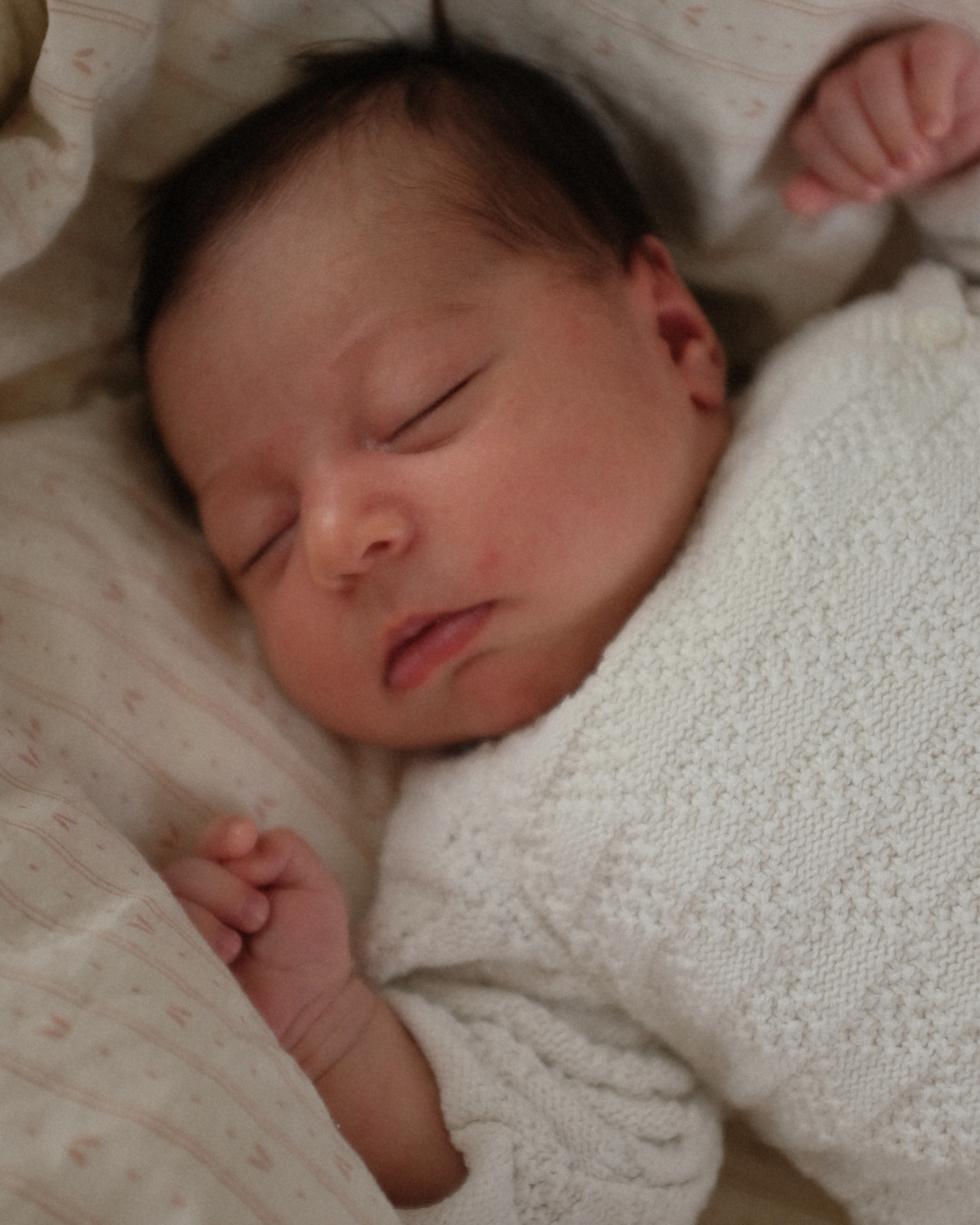 Close-up of a sleeping infant's face with closed eyes, lying on a patterned sheet, partially covered by a white blanket, showing soft skin and dark hair.