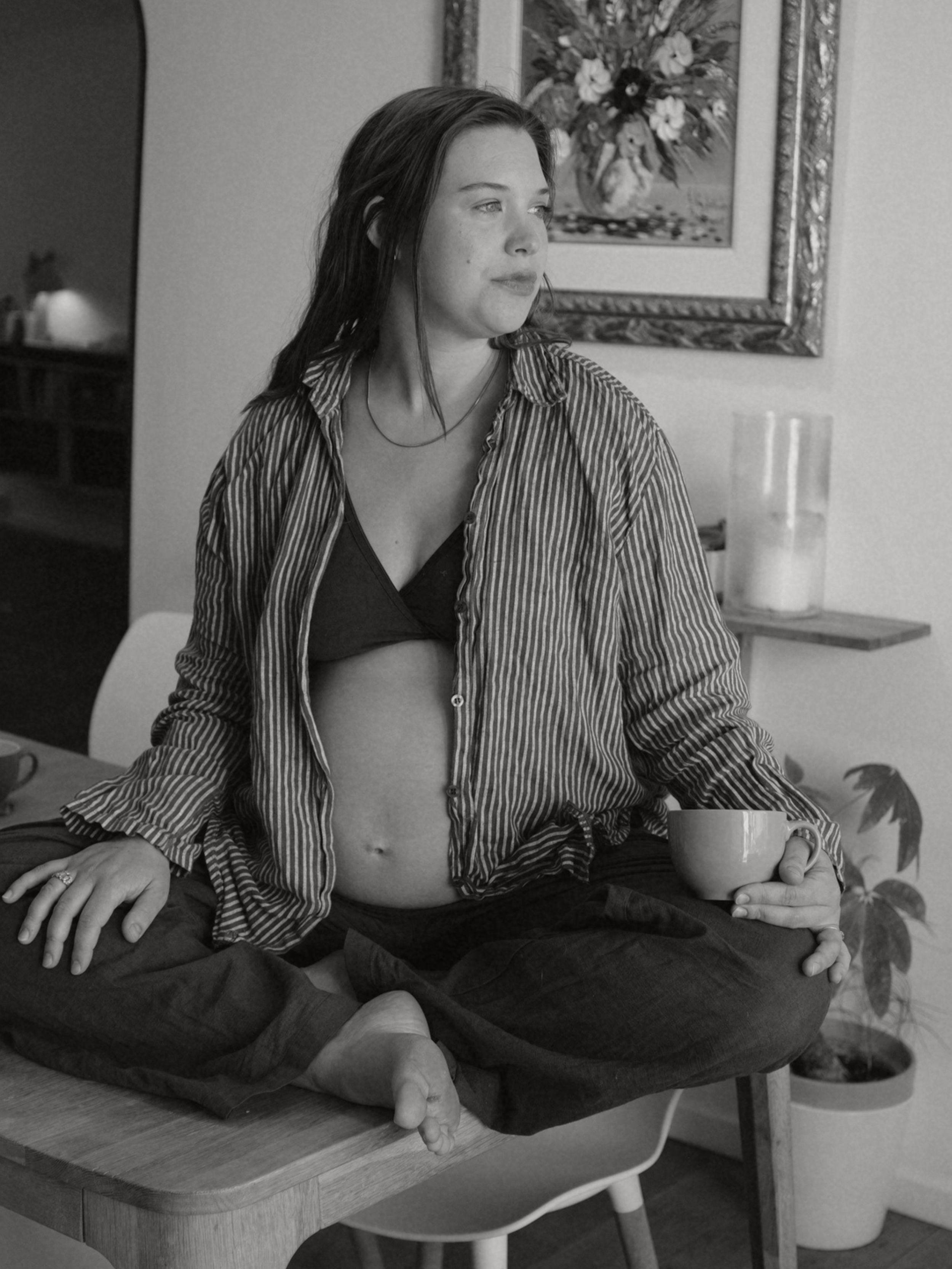 A woman with long dark hair, wearing a striped shirt and black bra, is sitting cross-legged on a table, holding a mug in her hand, in a room with a painting on the wall and a candle on a shelf.