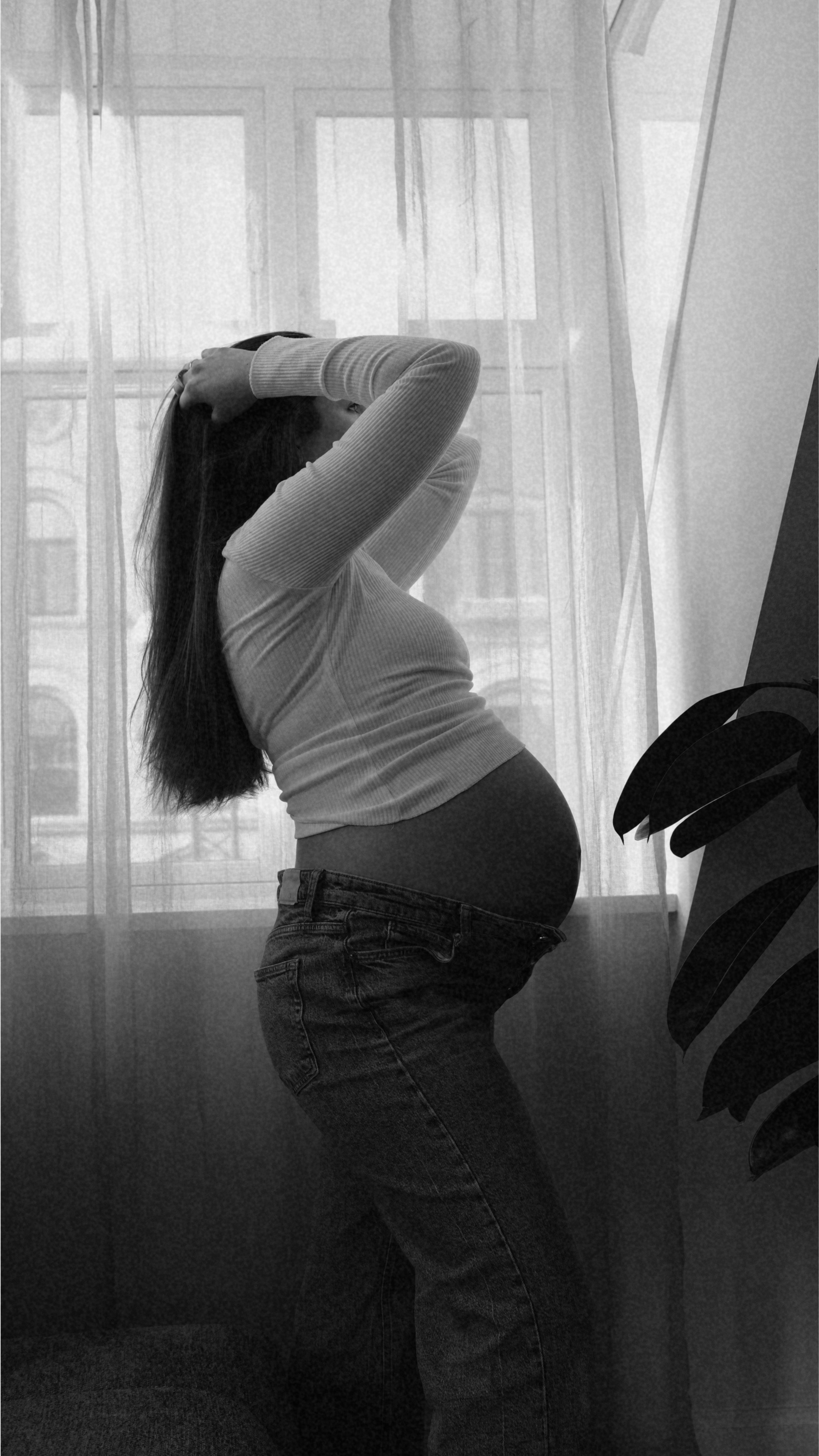 A pregnant woman standing in front of a window with sheer curtains, wearing a light-colored long-sleeve shirt and jeans, with her hands on her head. There is a plant with large leaves partially visible in the foreground.