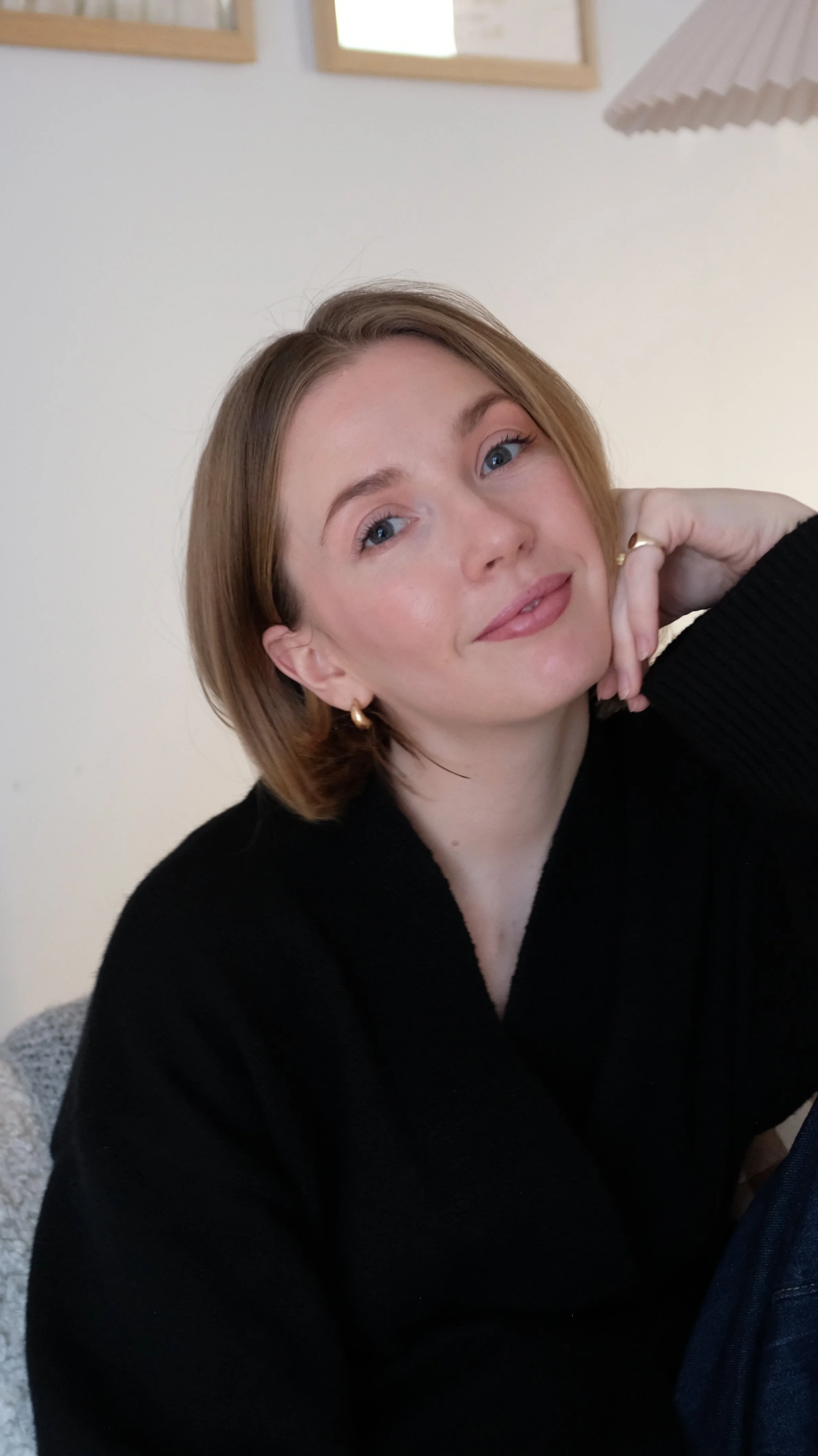 A woman with short blonde hair and blue eyes, wearing a black sweater and gold hoop earrings, resting her chin on her hand and smiling gently at the camera. The background shows part of a wall with two wooden frames and a white paper lampshade.
