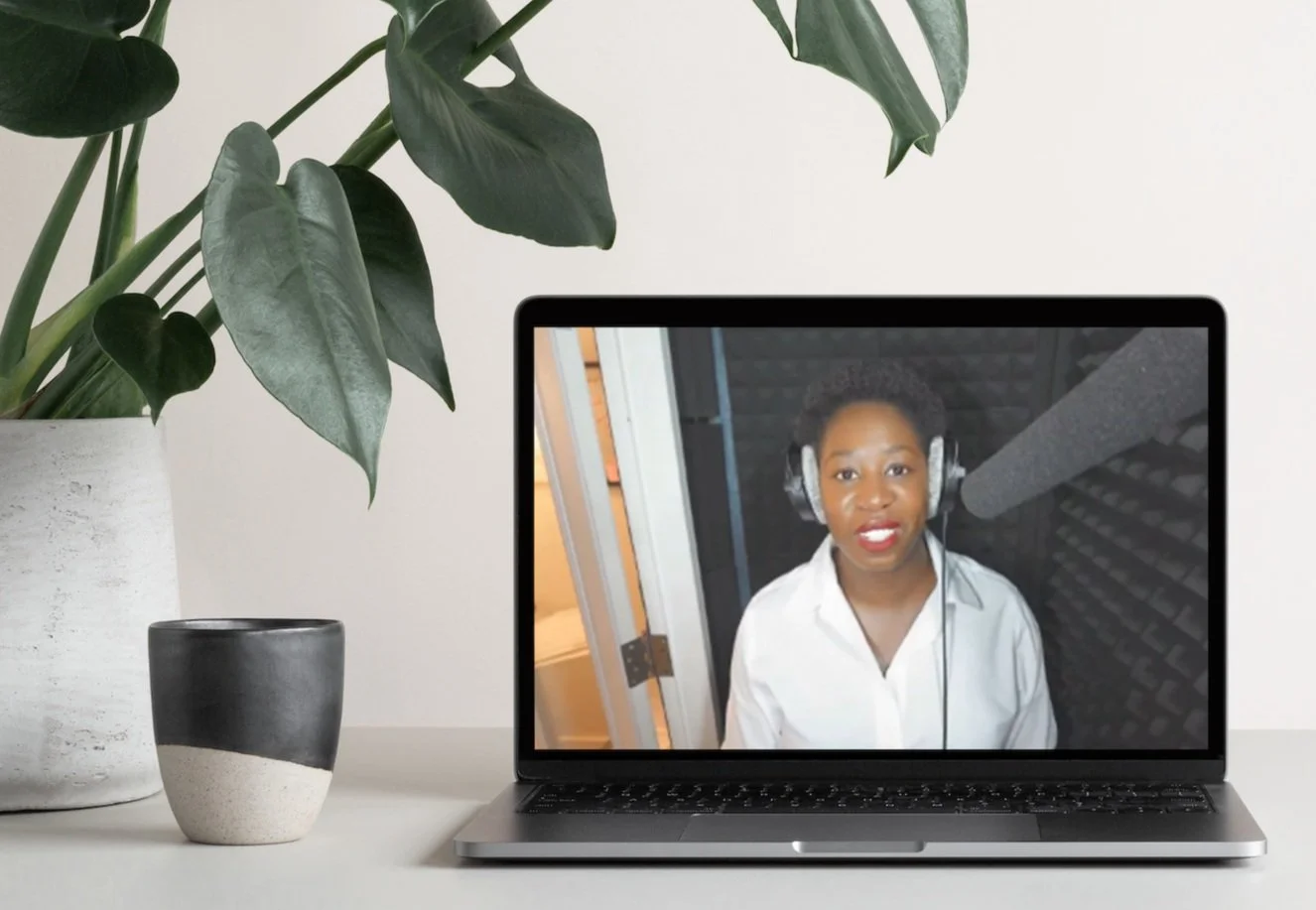 Laptop displaying a person wearing headphones in a recording studio, with a mug and potted plant nearby. This is Danielle Famble - a BusinessOPS client.