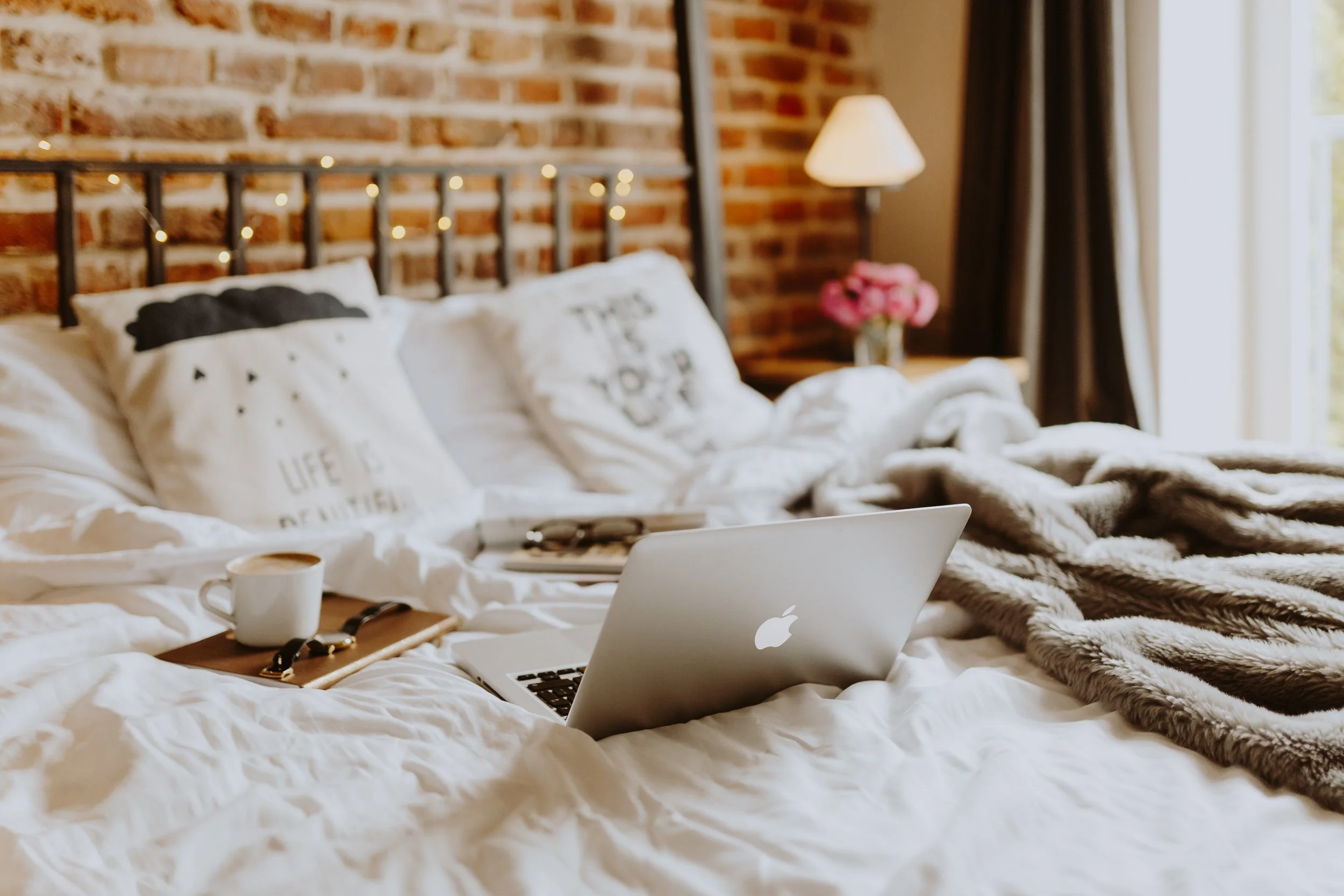 Cozy bedroom with a white bed, pillows, a faux fur blanket, and an open laptop. There is a coffee cup, a watch, and a notepad on the bed. A brick wall with string lights, a nightstand with a lamp and pink flowers, and a window with curtains are in th