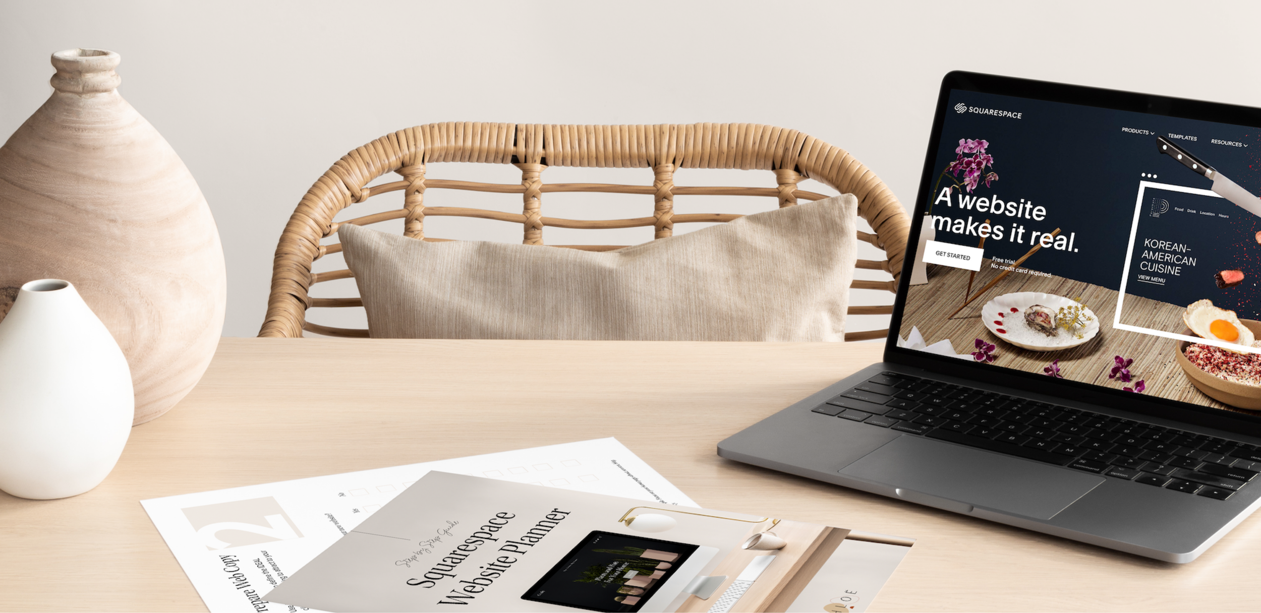 A modern workspace with a laptop displaying a website, a wicker chair with a beige pillow, two ceramic vases, and printed documents on a light wooden table.