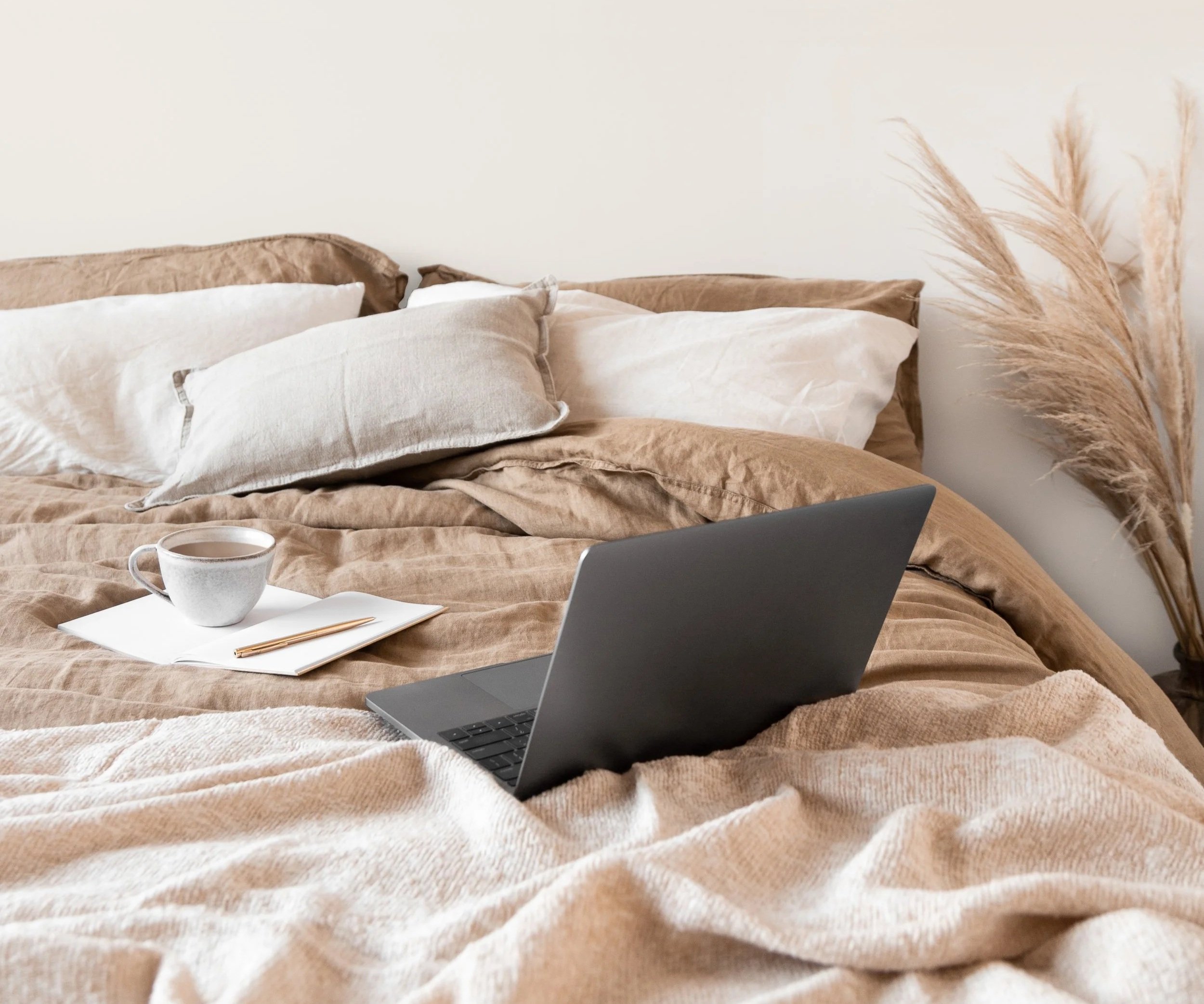 A cozy bed with beige and white pillows, a laptop, a cup of coffee, and some papers with a pen, in a minimalist bedroom with dried plants in the background.