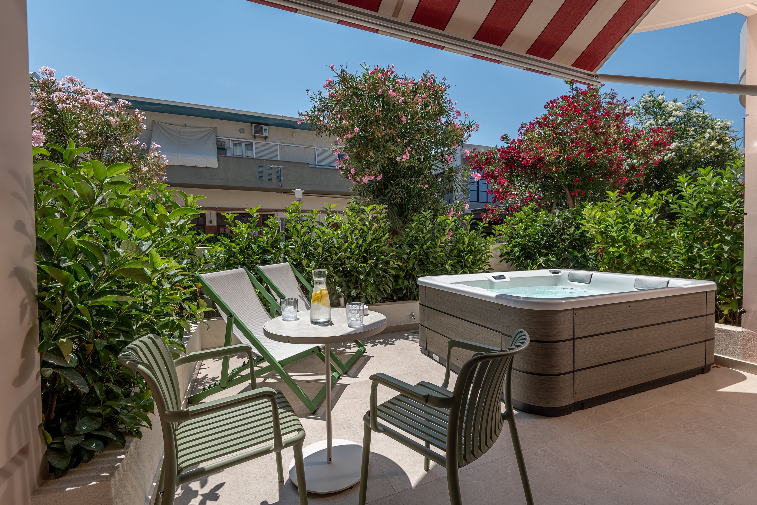 A patio with green chairs around a round table that has glasses and a bottle, a hot tub, lush bushes and flowering trees, and a building with balconies in the background under a clear blue sky.