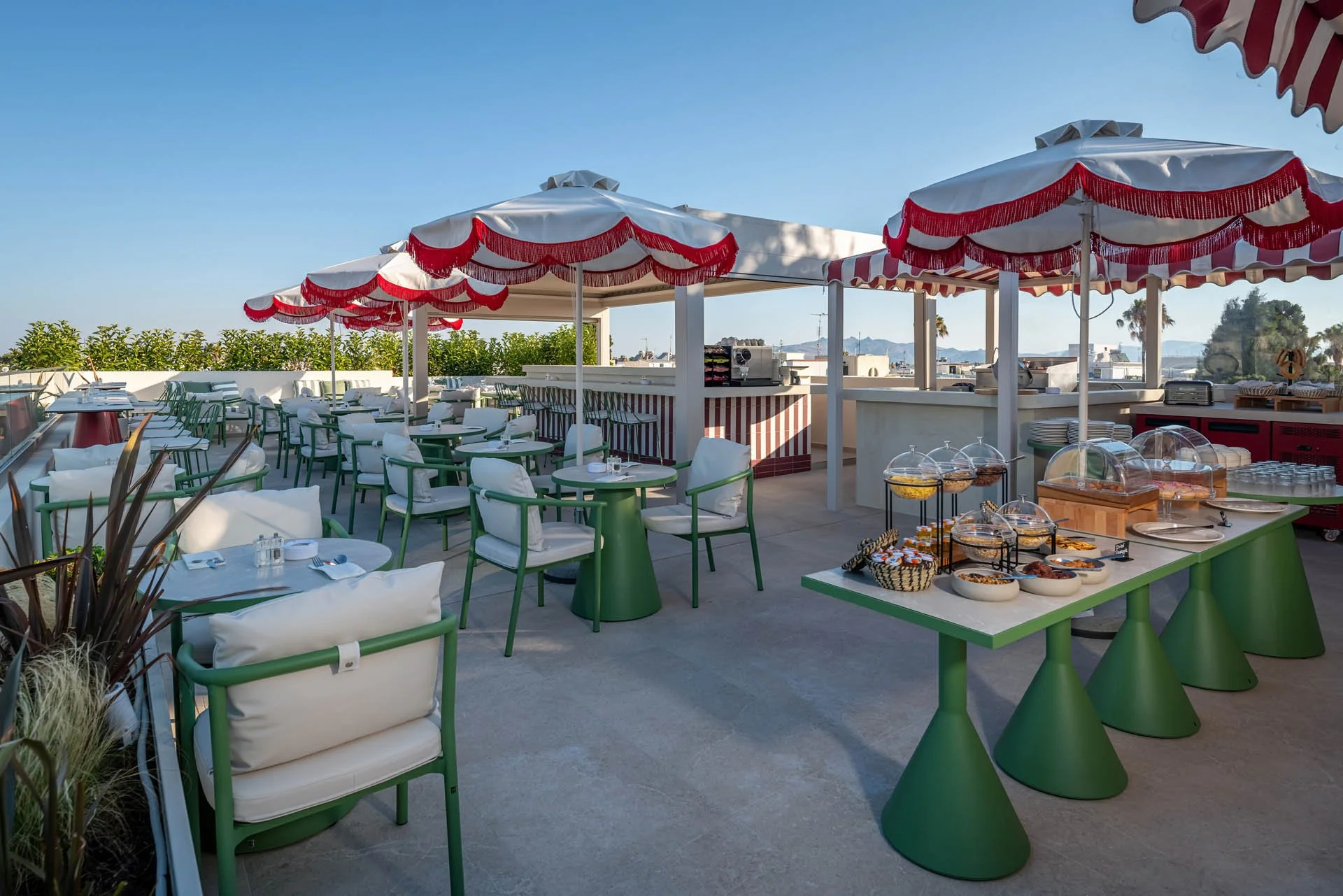 Outdoor restaurant patio with green chairs and white cushions under red and white striped umbrellas. There is a buffet table with food and plates on the right side. In the background, there are trees, shrubs, and a clear blue sky.