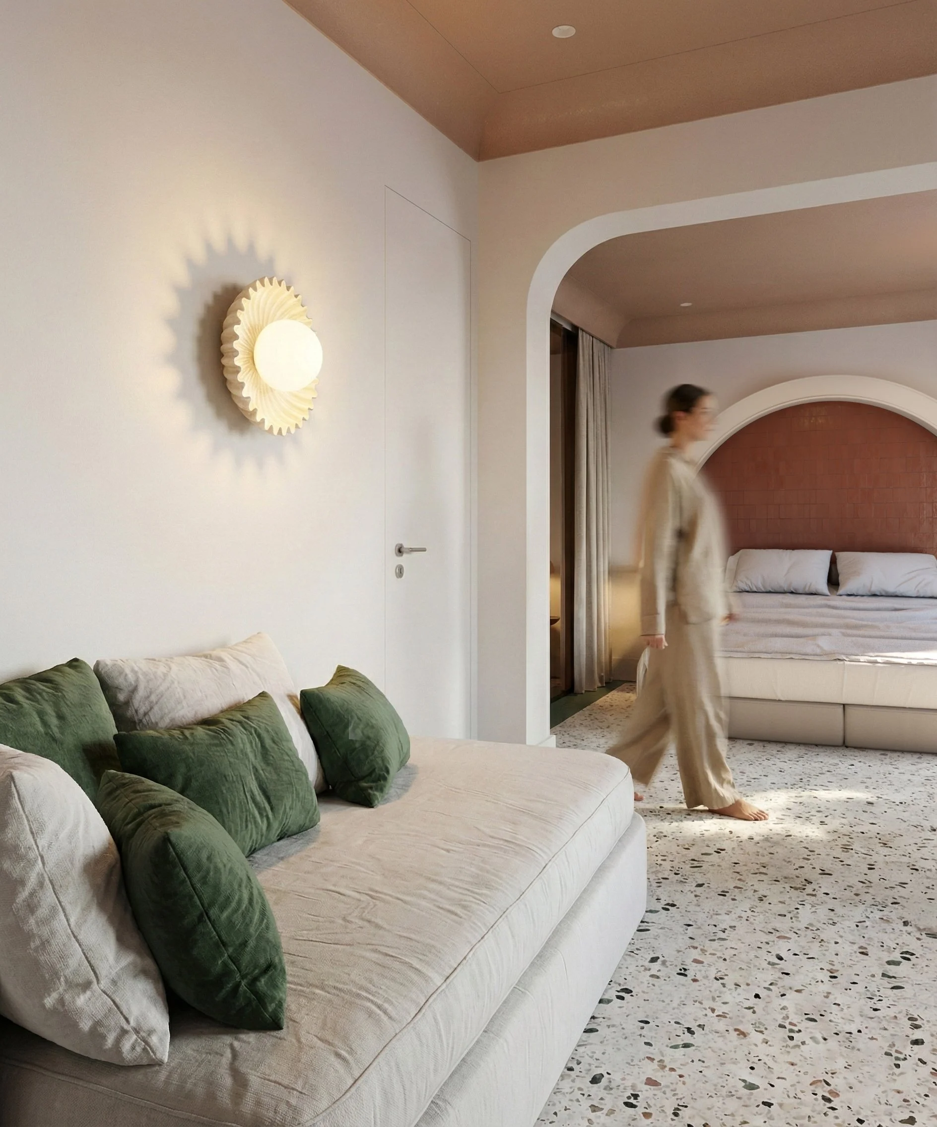 Modern hotel room with a white sofa and green pillows, a woman in beige pajamas walking, terrazzo flooring, and a bed with a brick red headboard.
