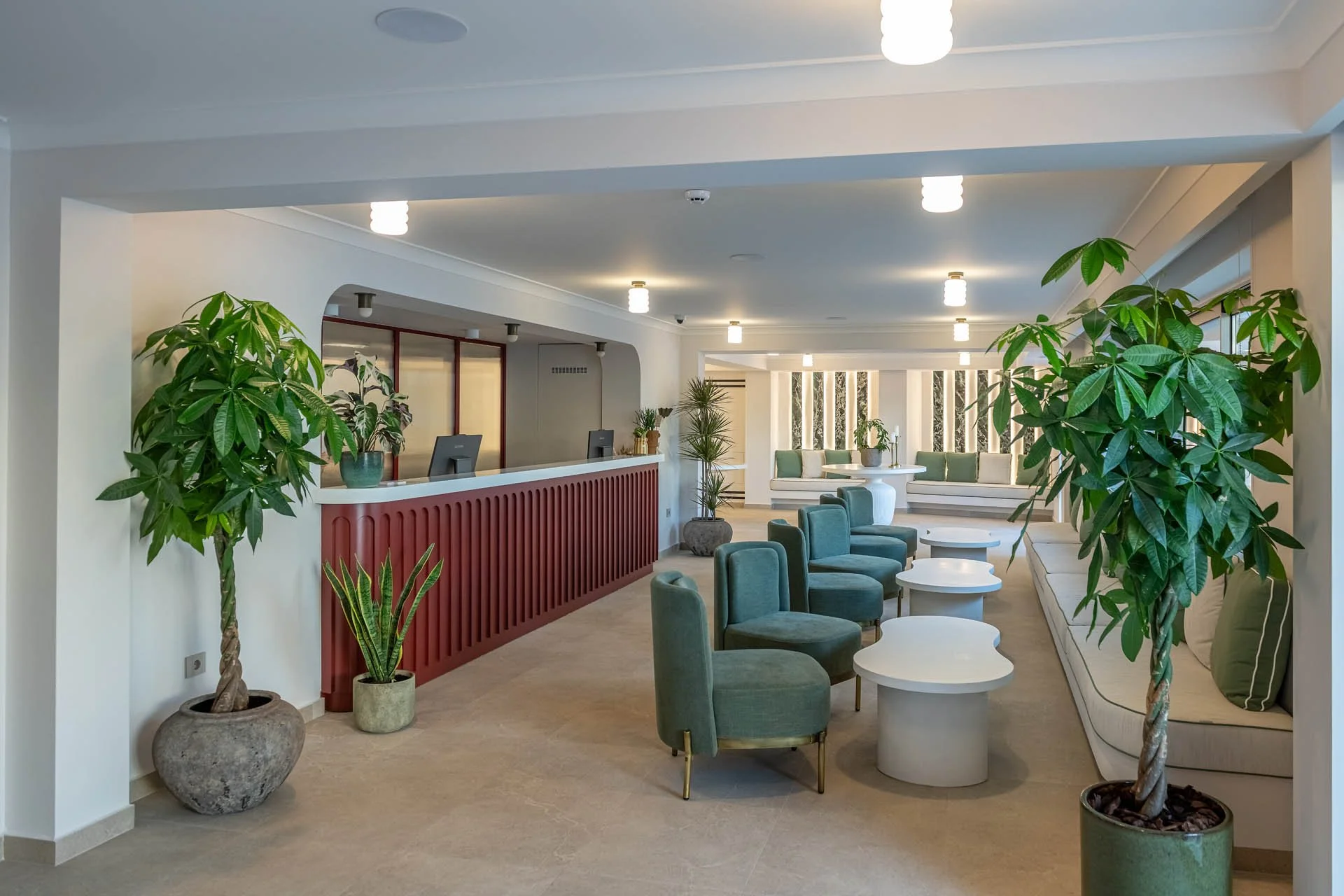 A spacious modern lounge area with green armchairs, round white tables, and potted green plants, including palm-like plants, near the reception desk with computers. Large windows with vertical striped curtains in the background, ceiling lights, and neutral-colored walls and flooring.