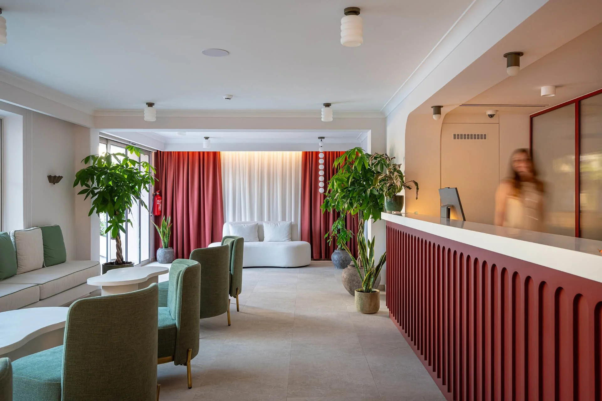 Hotel lobby with seating area, large potted plants, and reception desk with a blurred woman in the background.