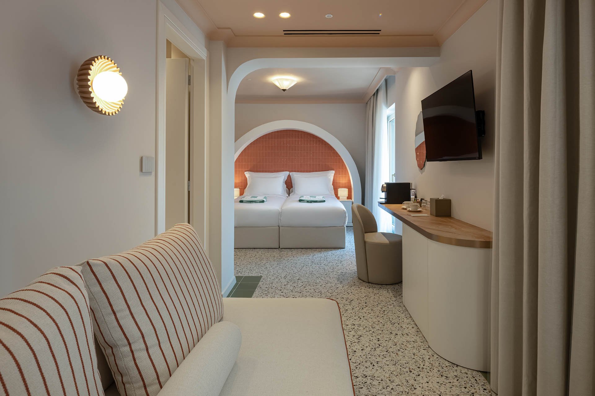 View of a hotel room with a sitting area, a desk, and a bedroom with two beds, orange headboards, and white linens. The room has a modern design with light-colored walls, ceiling lights, a wall-mounted TV, curtains, and a terrazzo floor.