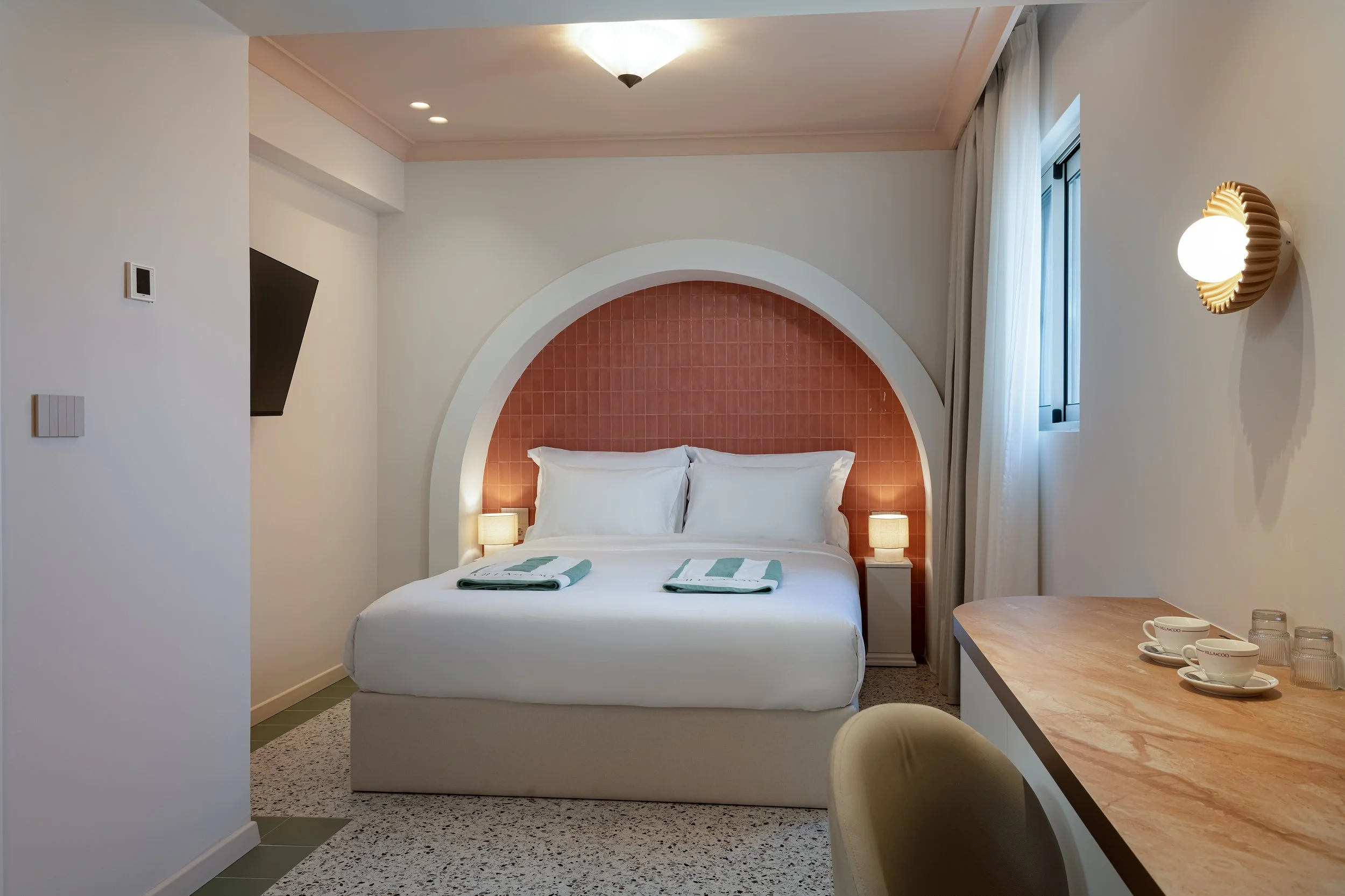 A hotel room with a bed, two lamps, a pink-tiled headboard, and a window with white curtains.
