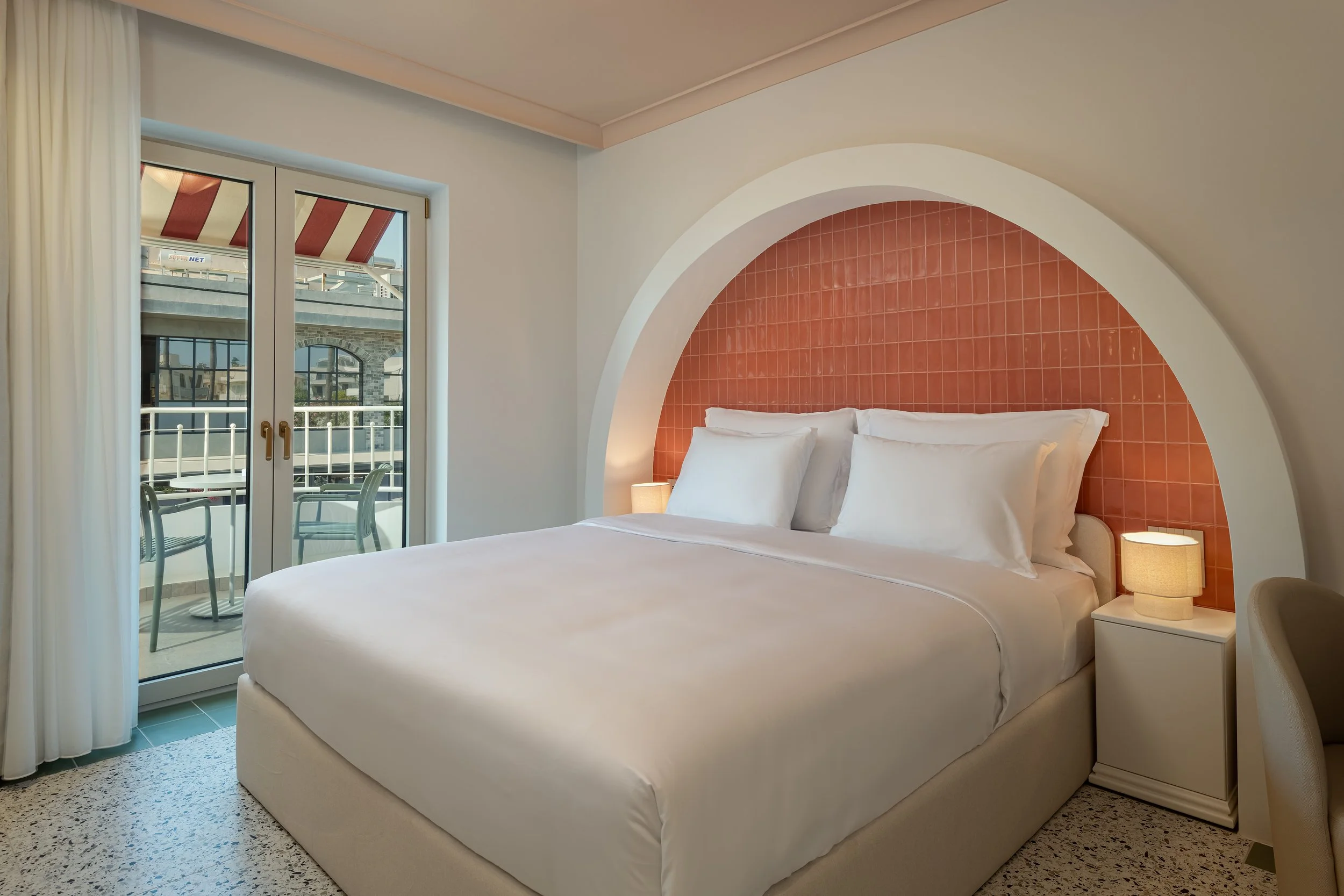 A modern hotel room with a white bed, bedside lamp, and a large window showing a balcony with chairs and an exterior building.
