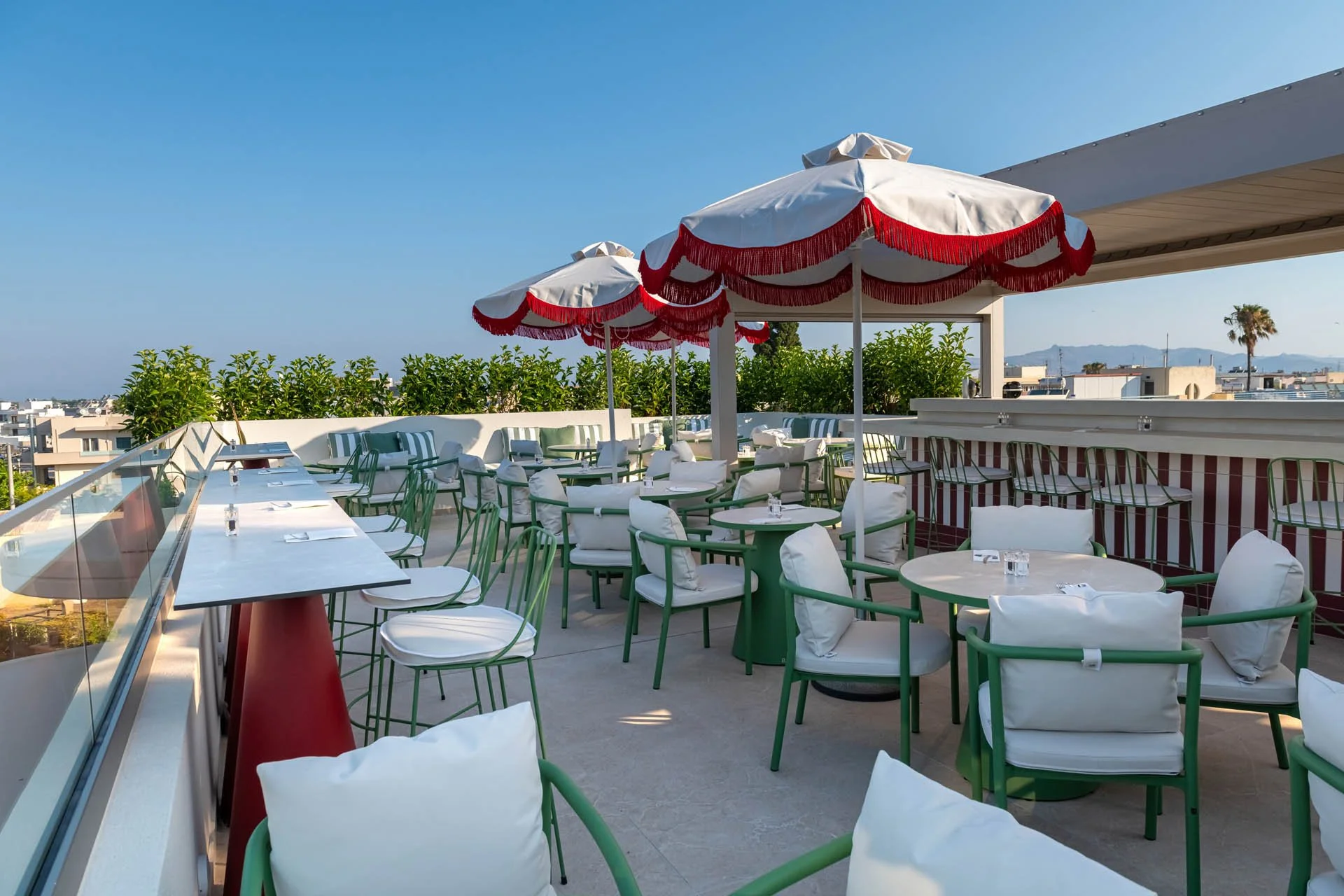 Rooftop restaurant or bar with white and green chairs, white cushions, large umbrellas trimmed with red fringe, set against a cityscape and mountain background under a clear blue sky.