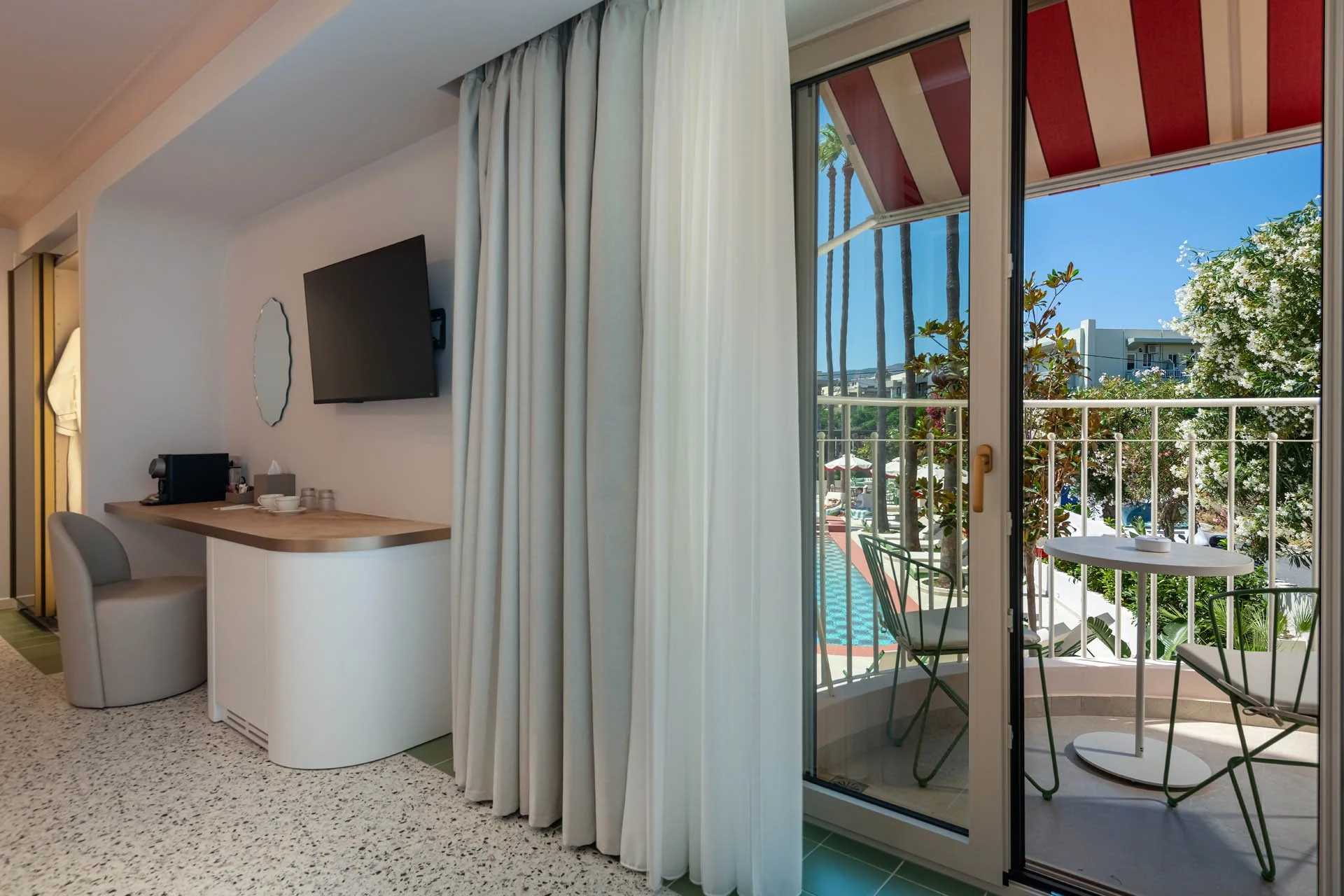 View of a hotel room's balcony with metal chairs and a small round table, with trees and buildings in the background, seen through an open sliding glass door with striped red and white awning outside.