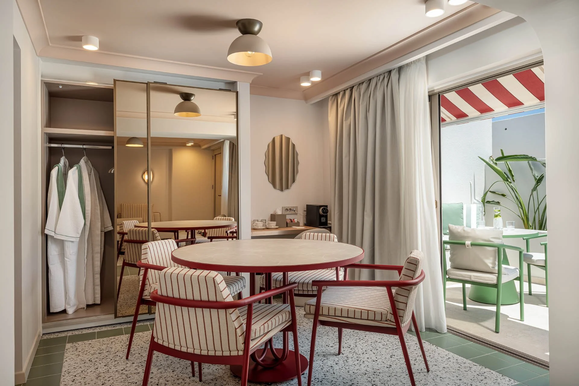 A cozy dining area with a round table surrounded by striped chairs, a mirror reflecting a small sitting space, open closet with robes, patio door with curtains, and a balcony with green chairs and a striped awning.