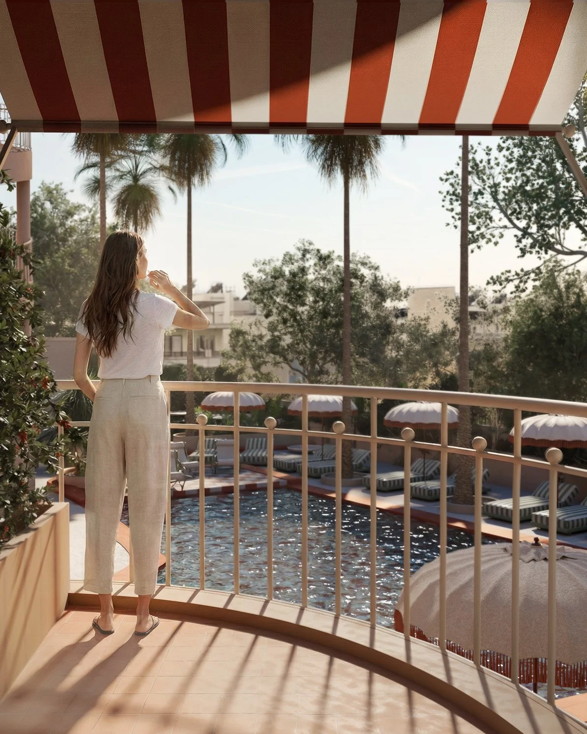 A woman in beige pants and a white top standing on a balcony overlooking a swimming pool with lounge chairs and umbrellas, with palm trees in the background.