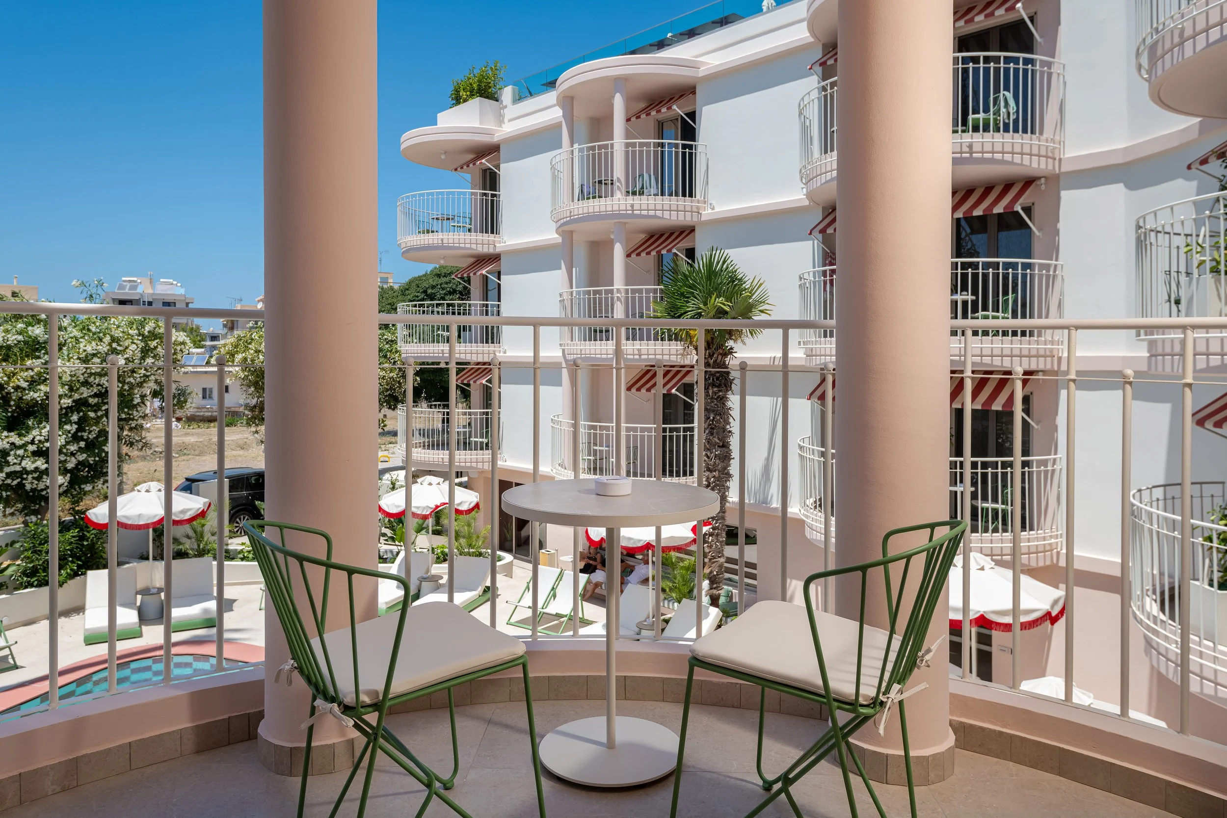 A balcony view of a modern hotel or apartment building with white curvy railings and pinkish-orange columns, green chairs around a white table, and a dense garden with white flowers and red umbrellas in the background, under clear blue sky.