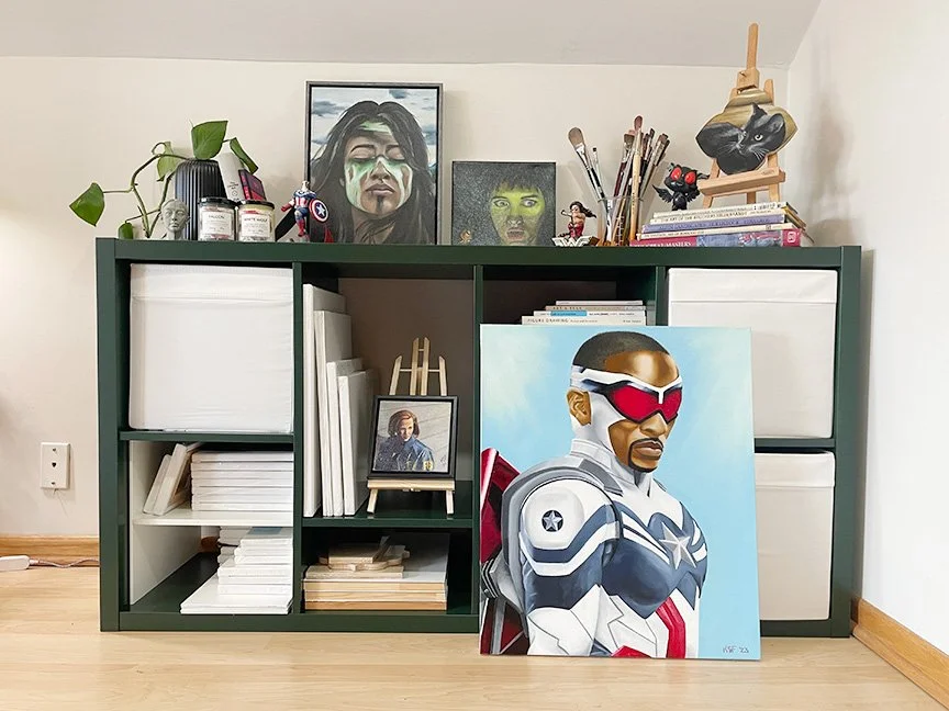 A green shelving unit with white storage bins, artwork, books, and art supplies on top. A large painting of a man in a futuristic suit with sunglasses leaning against the cabinet. Smaller art pieces and figurines are also on the top shelf.