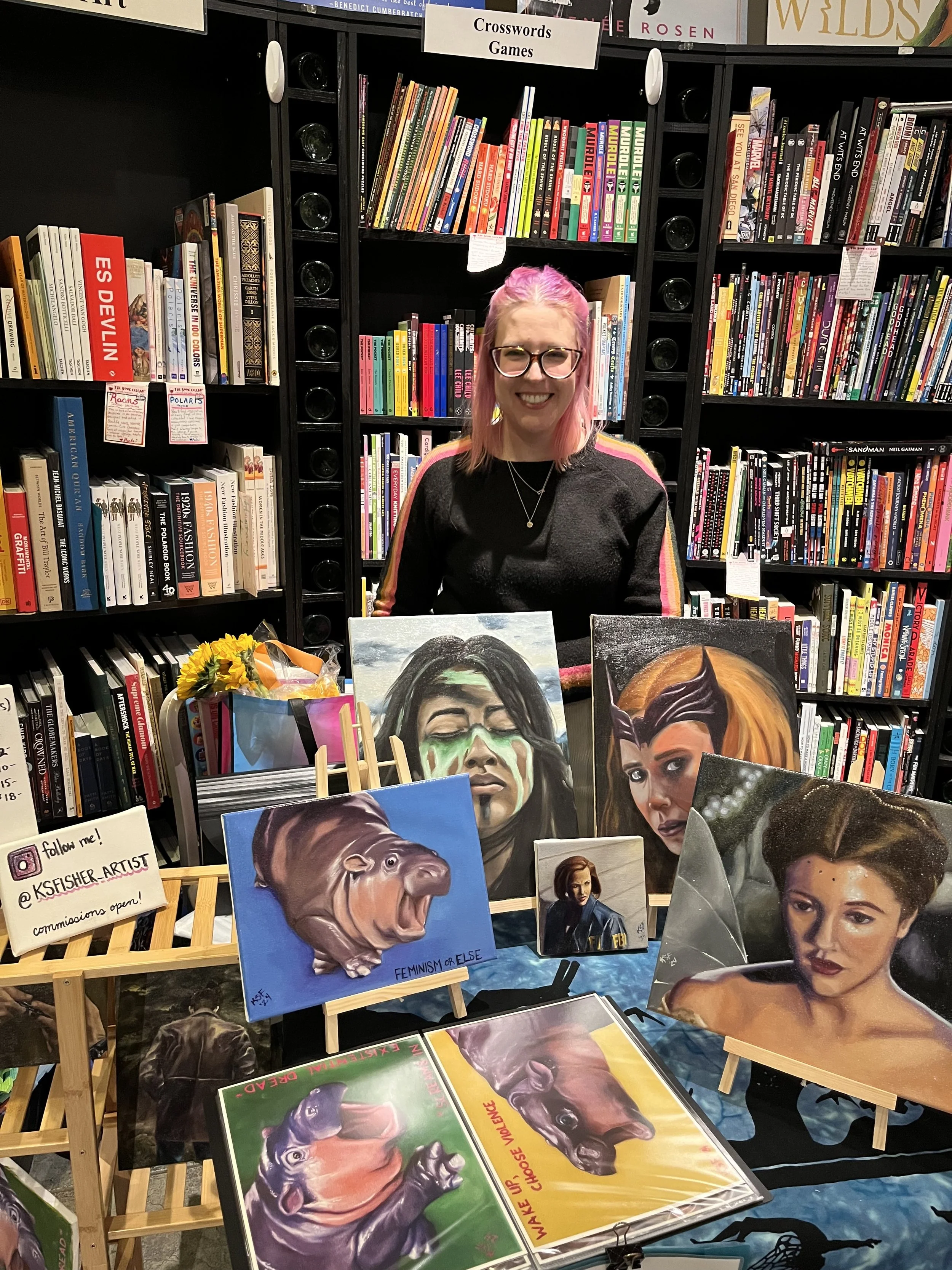 A woman with pink hair and glasses stands behind a table with various paintings of women and animals, at a bookstore or library. The background features bookshelves labeled 'Crosswords' and 'Games'.