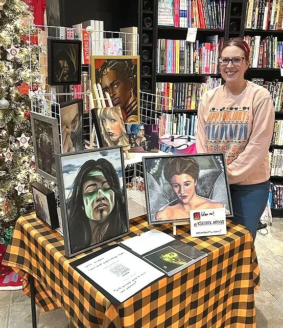 Woman standing behind a table with framed paintings of women, with a bookshelf behind her in a bookstore or library.
