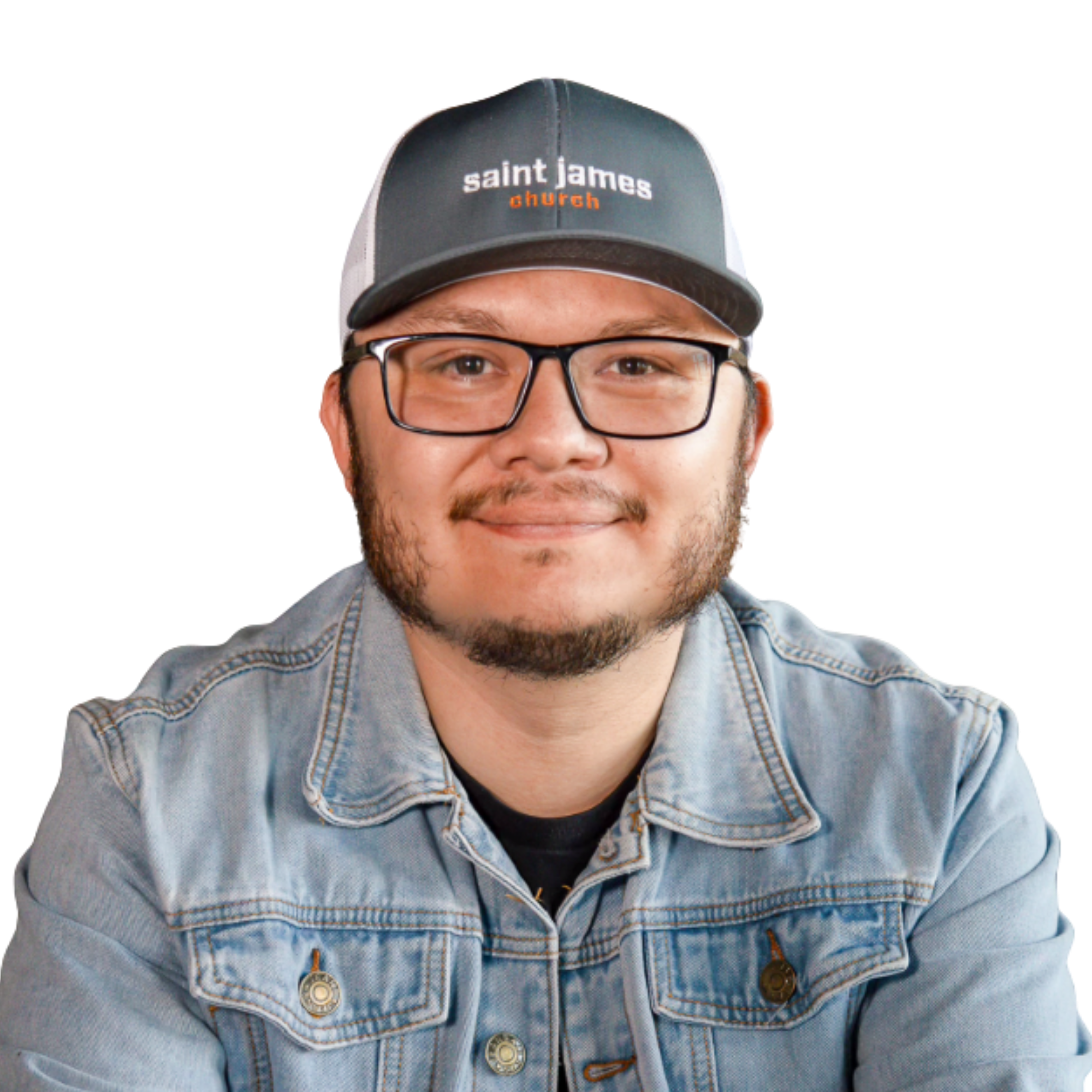 Close-up of a young man's face wearing glasses and a denim jacket, with a black and gray cap that has white and orange text reading 'saint james church'.