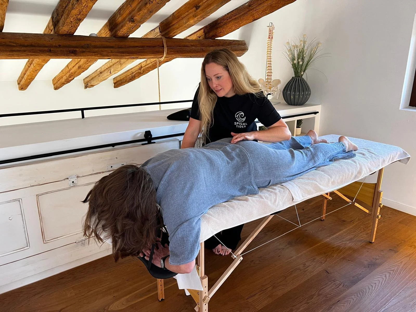 A woman giving a Spinal Flow Technique session to a person lying face down on a massage table in a room with wooden beams and white walls.