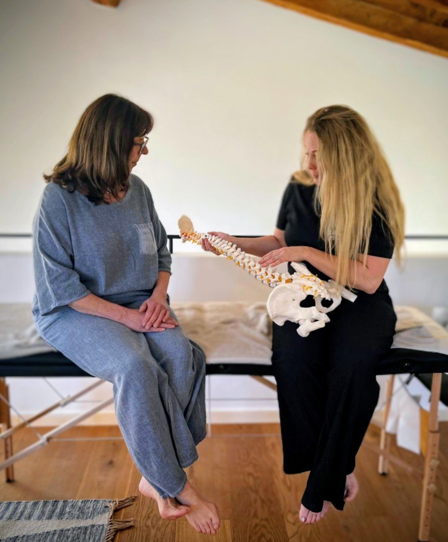 Two women sitting on a massage table, with one woman showing the other a model of the human spine and pelvis, in a room with wooden floors and walls.
