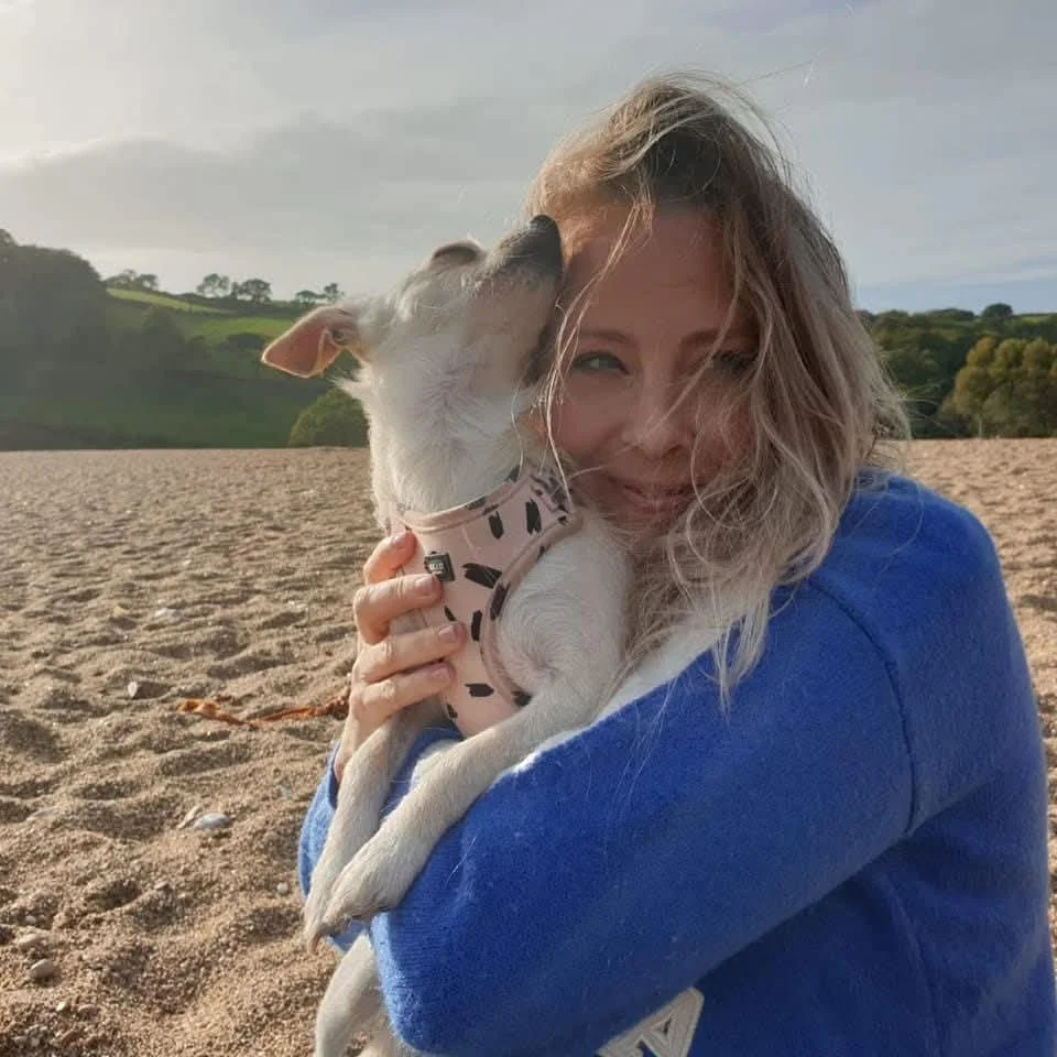 A woman with long, curly blonde hair smiling while holding a small dog on a sandy beach. The dog is licking her face and wearing a pink harness. Hills and trees are visible in the background.
