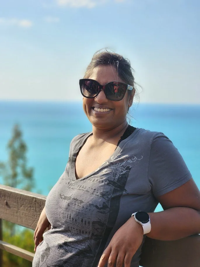 Woman wearing sunglasses and a gray t-shirt, smiling, standing outdoors near a wooden railing with a scenic view of the ocean and sky in the background.