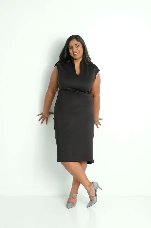 A woman with dark hair in a black dress and gray high heels standing against a plain white background.