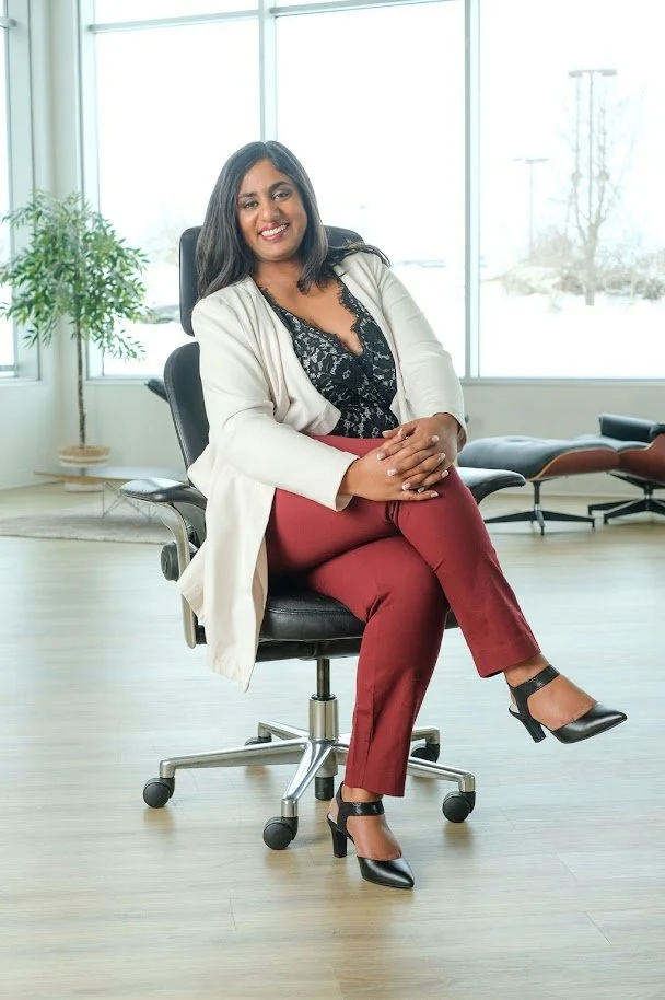 A woman sitting on an office chair in a modern office with large windows, a potted plant, and co-working chairs in the background, smiling and dressed professionally.