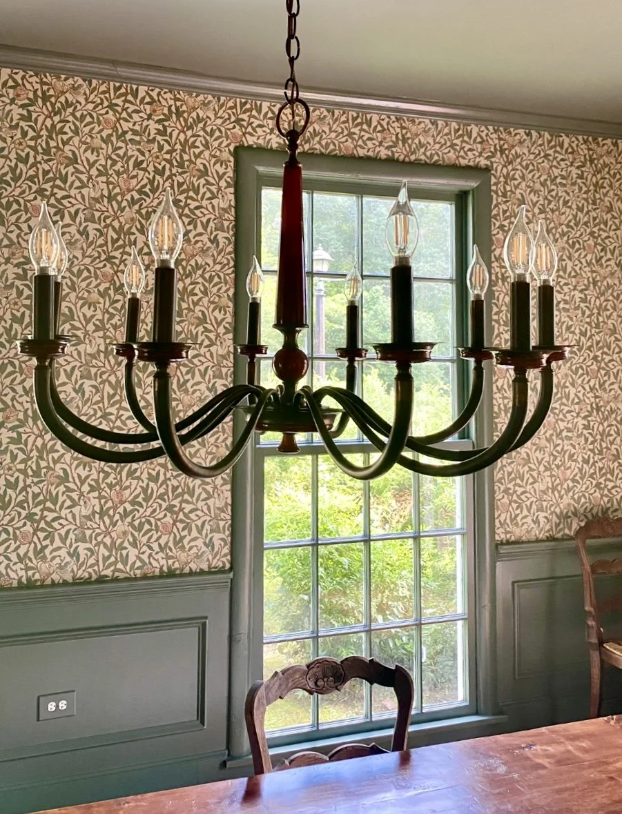 A black chandelier with candle-shaped light bulbs hangs above a wooden dining table in a room with floral wallpaper and a large window.