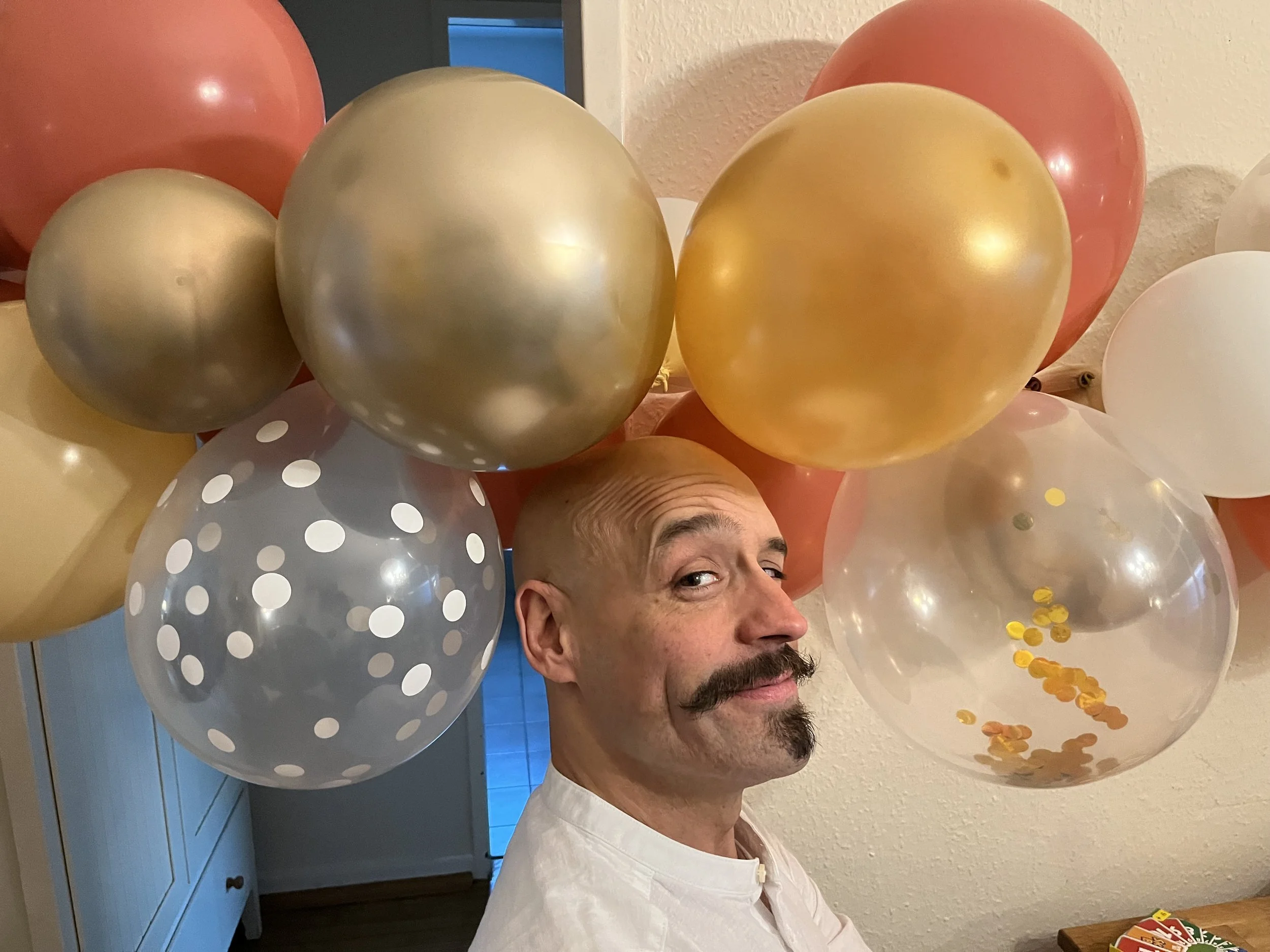 Ein Mann mit Schnurrbart und Glatze liegt im Raum, umgeben von vielen Ballons in Gold, Weiß, Rosé und transparent mit Konfetti, die an der Decke hängen oder gehalten werden.