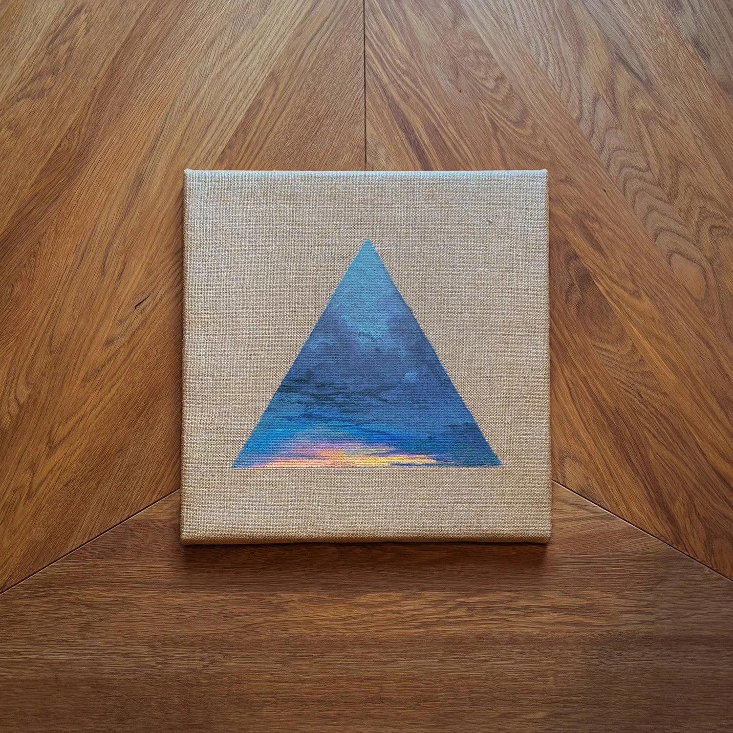 Kleines quadratisches Bild mit einem blauen Dreieck, das eine Himmelsszene mit Wolken und Sonnenuntergang zeigt, auf einer beige Leinwand, auf einem Holztisch