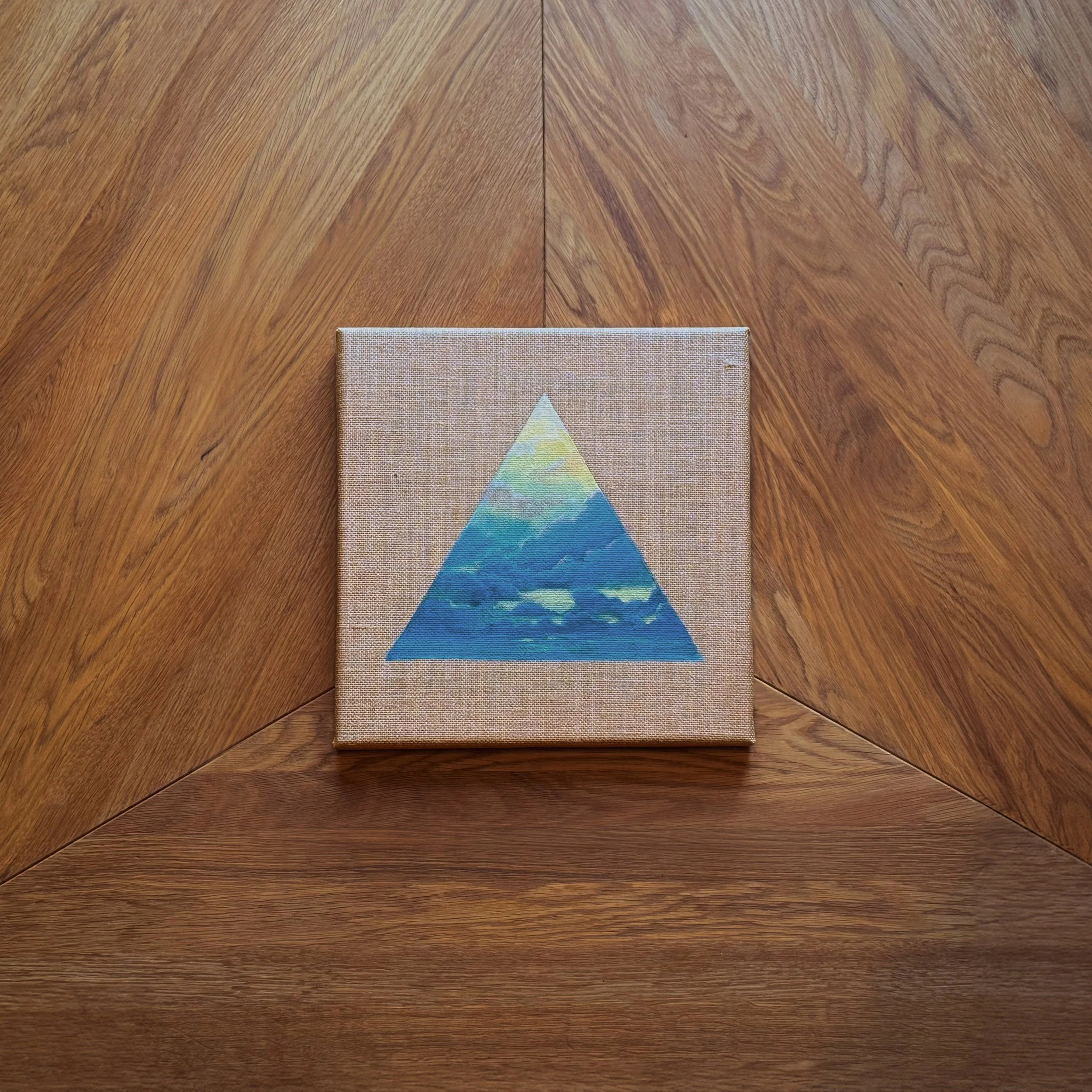 Kleines Gemälde auf Leinwand mit einem blauen, dreieckigen Muster, das einen Himmel mit Cloud- und Sonnen-Elementen darstellt, auf einem Holzfußboden platziert.